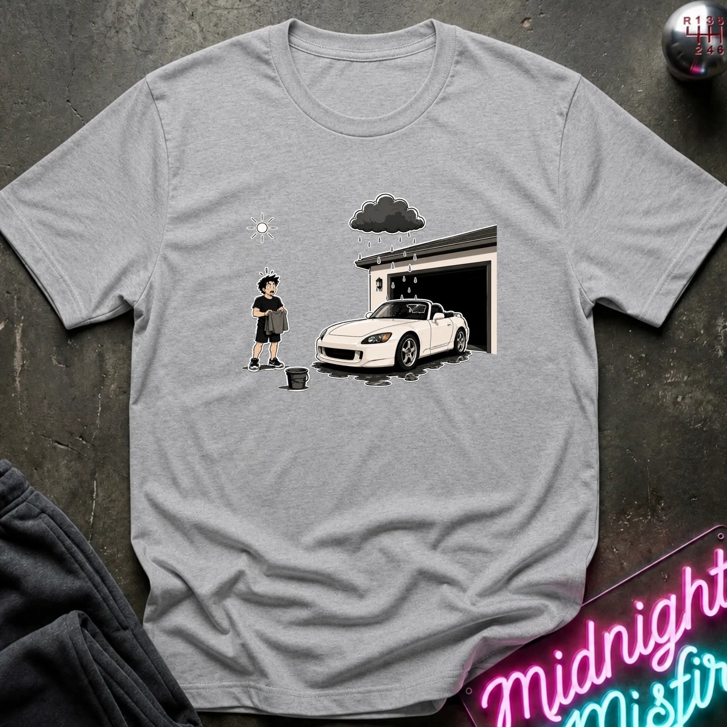 A heather gray short-sleeve t-shirt with a graphic print of a person washing a white sports car in front of a garage under a dark cloud. The t-shirt is laid flat on a dark textured surface with a neon sign and dark pants partially visible.