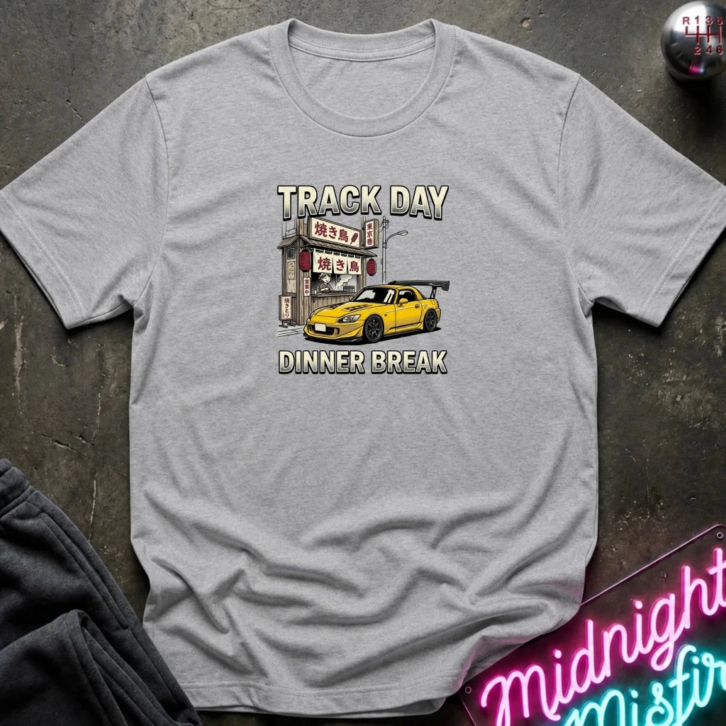 A light gray heather crewneck t-shirt featuring a graphic print of a yellow sports car parked in front of a Japanese street sign with the text 'TRACK DAY DINNER BREAK'. The shirt is laid flat on a dark textured surface with a neon sign and dark pants partially visible in the corners.