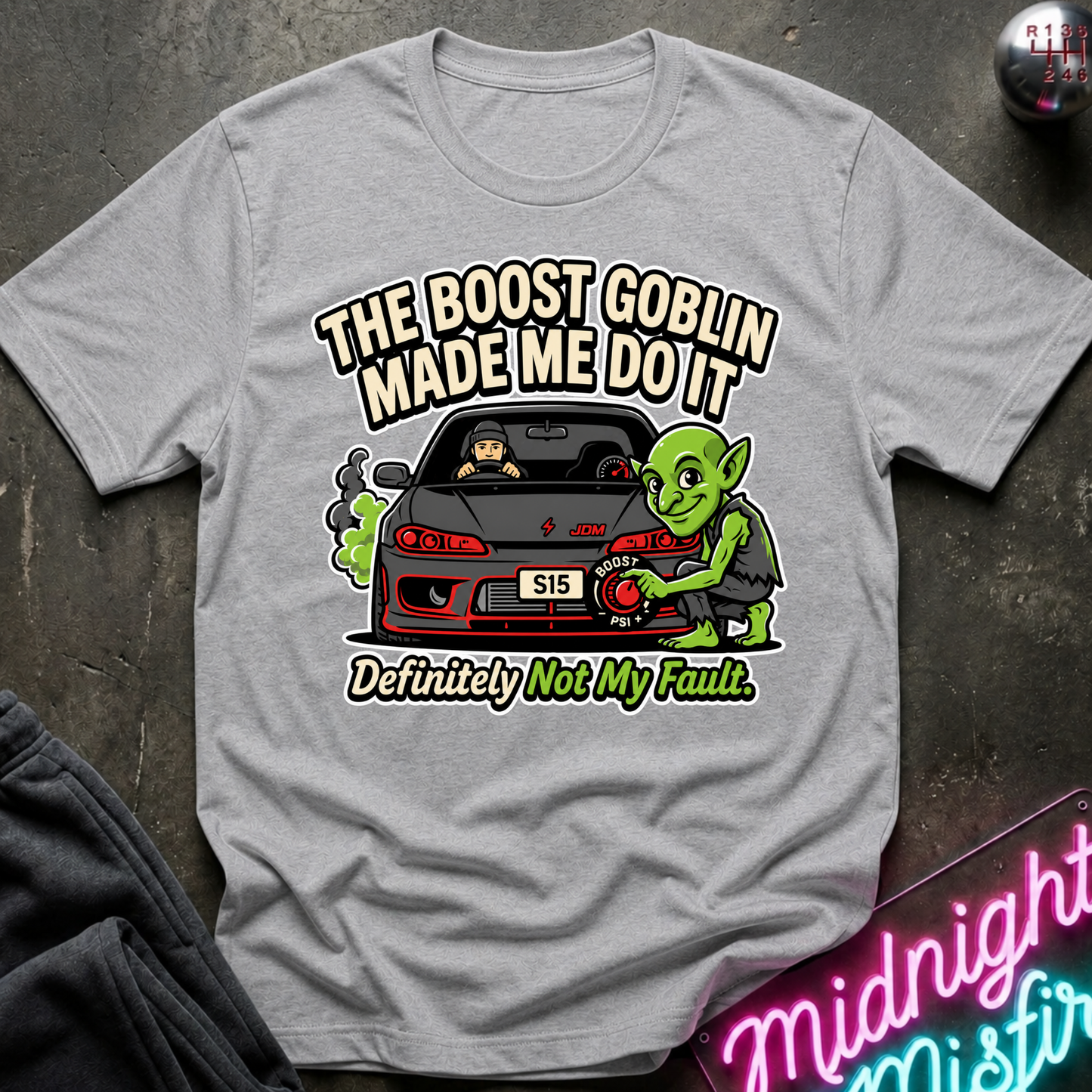 A light gray graphic t-shirt laid flat on a dark textured surface. The shirt features a cartoon illustration of a green goblin fixing a black sports car with the text 'THE BOOST GOBLIN MADE ME DO IT' and 'Definitely Not My Fault.