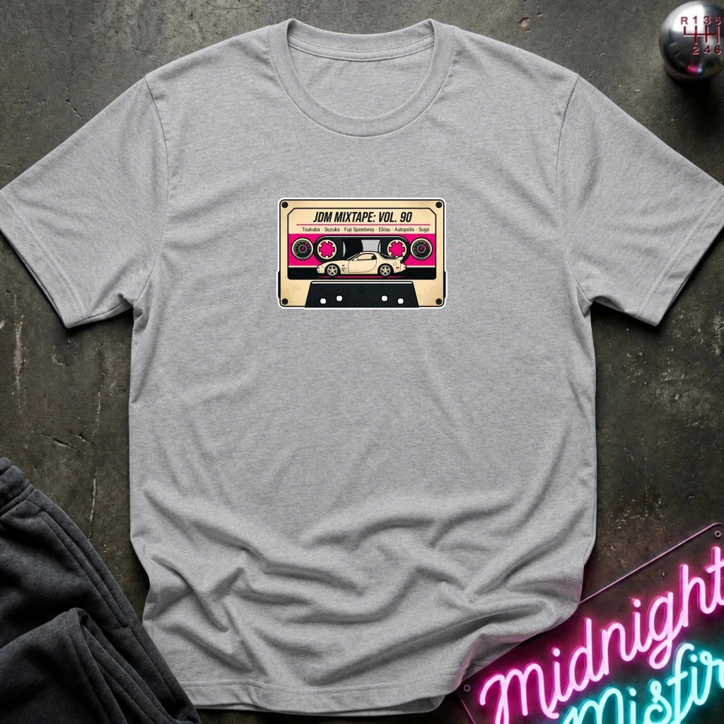 A heather gray crewneck t-shirt laid flat on a dark textured surface, featuring a retro cassette tape graphic on the chest with the text 'JDM MIXTAPE VOL.
