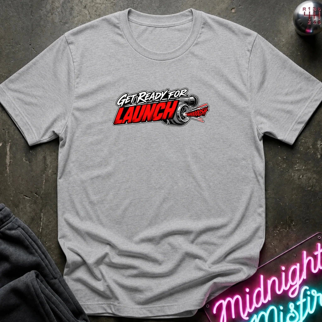 A heather gray short-sleeve t-shirt laid flat on a dark textured surface, featuring a 'GET READY FOR LAUNCH' graphic with a red and black rocket design across the chest.