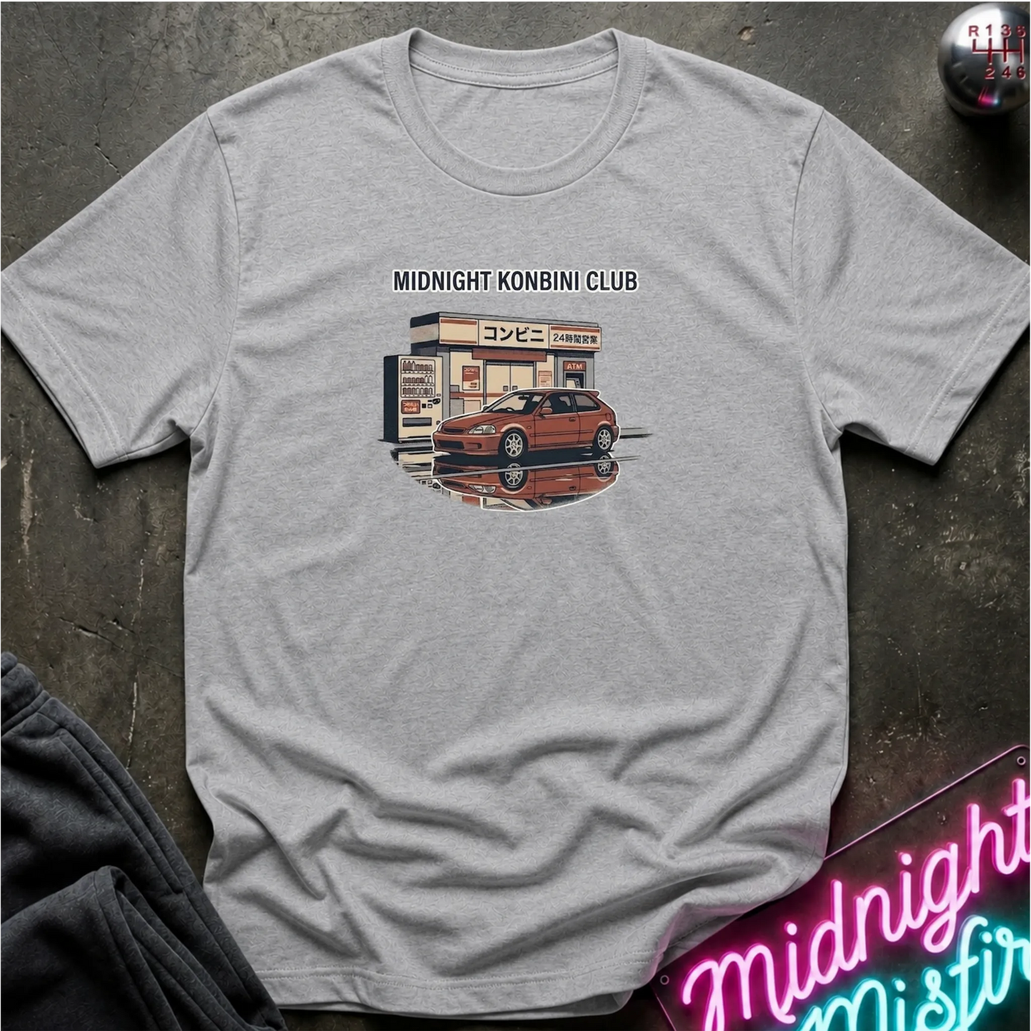 A light gray heather crewneck t-shirt laid flat on a dark textured surface. The shirt features a graphic print of a red sports car parked in front of a convenience store with the text 'MIDNIGHT KONBINI CLUB' above it.