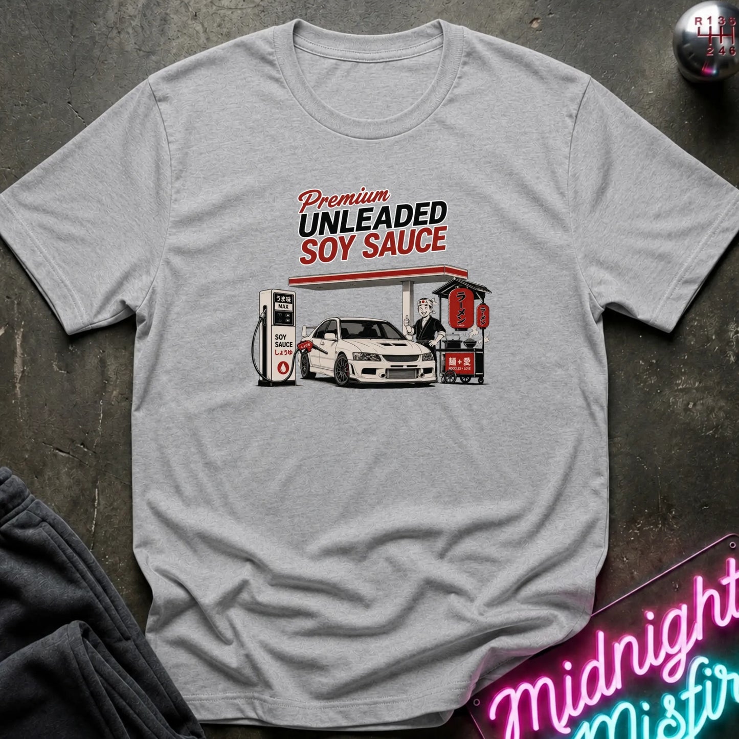 A light gray heather crewneck t-shirt featuring a retro-style graphic of a gas station pump and car with the text 'Premium UNLEADED SOY SAUCE'. The shirt is laid flat on a dark textured surface with a neon sign and dark pants partially visible in the corners.