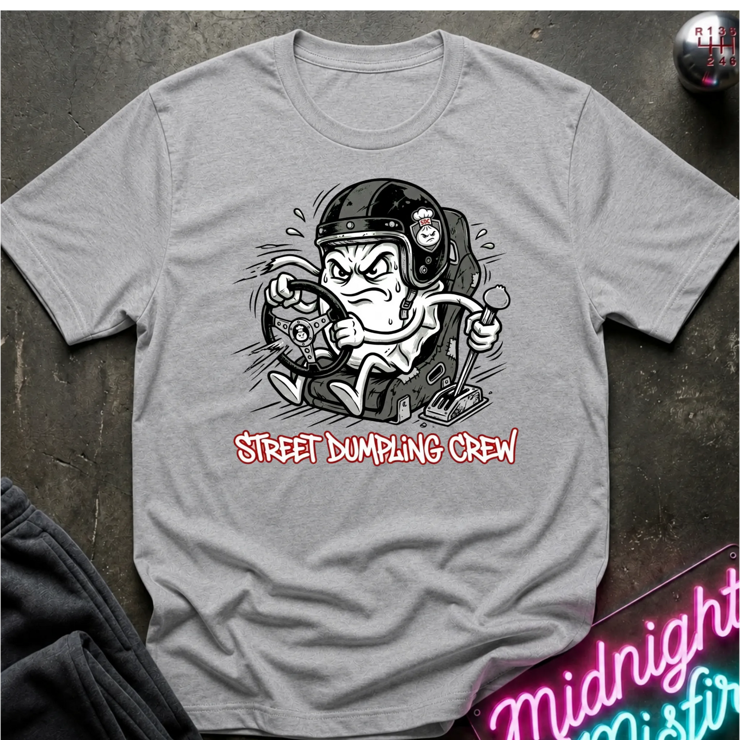 A heather gray graphic t-shirt laid flat on a dark textured surface. The shirt features a cartoon illustration of a street-dumping character in a helmet with the text 'STREET DUMPING CREW' printed below in red and white.