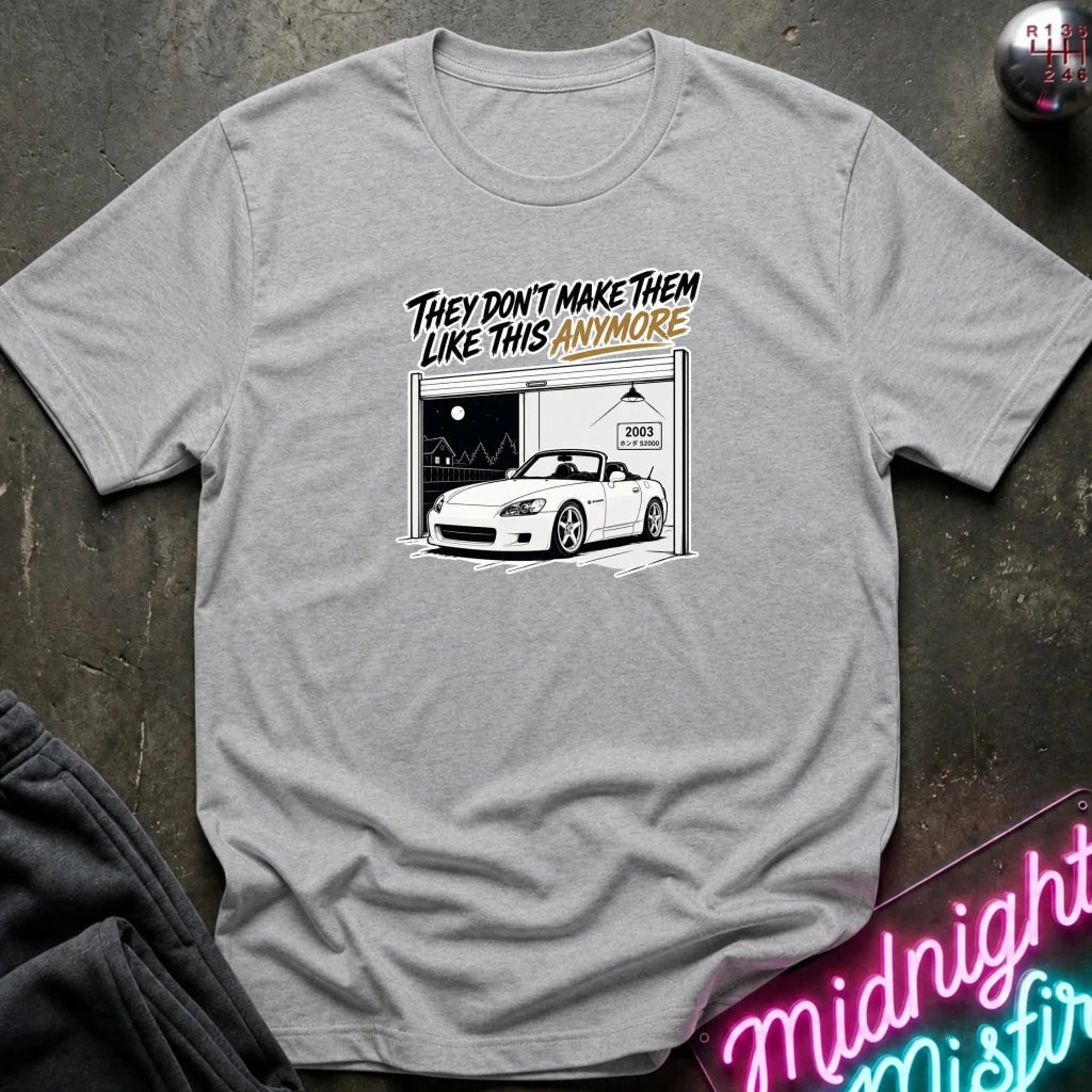 A heather gray short-sleeve t-shirt laid flat on a dark textured surface. The shirt features a graphic print of a white sports car parked inside a garage with the text 'THEY DON'T MAKE THEM LIKE THIS ANYMORE' in black and orange lettering.