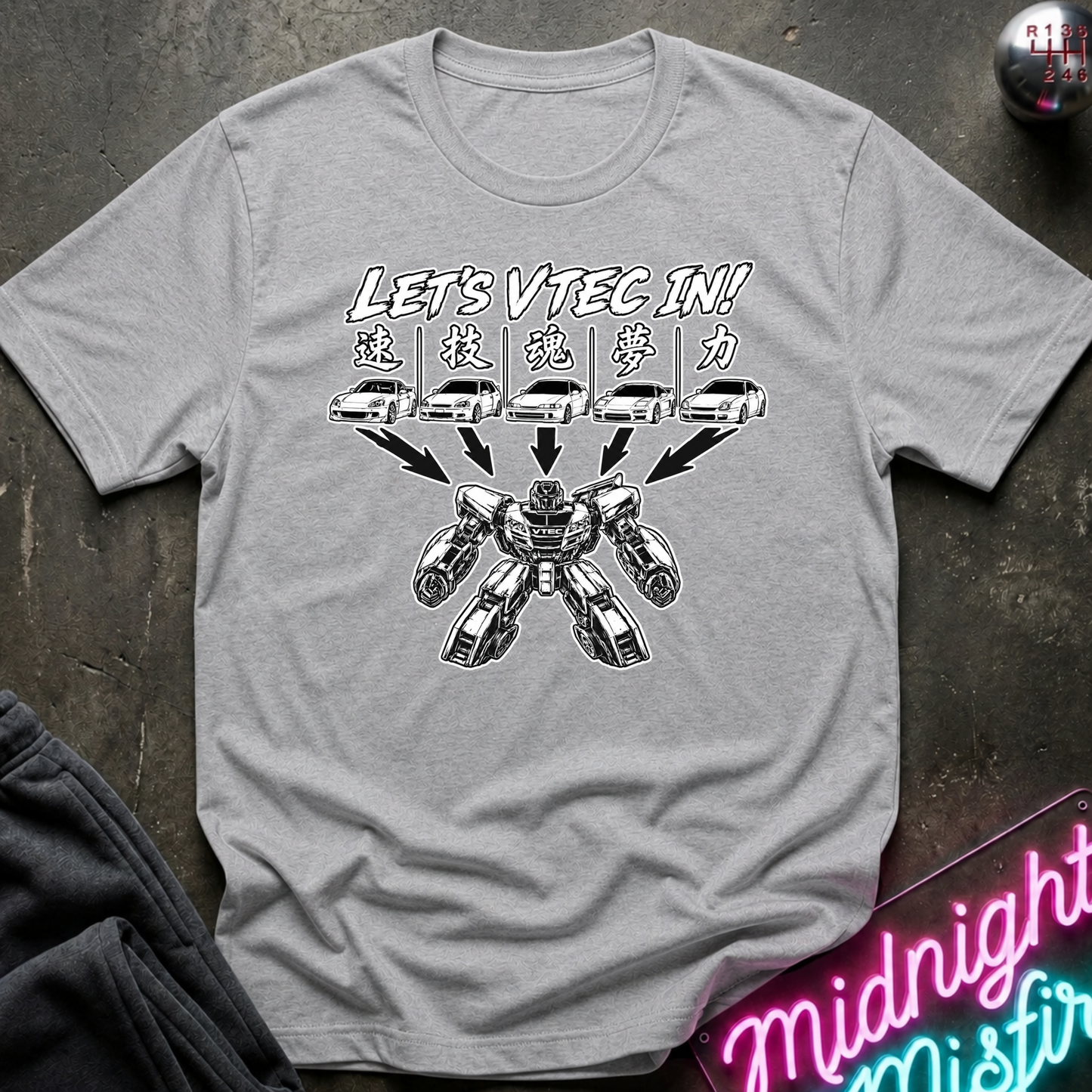 A heather gray short-sleeve t-shirt with a black and white graphic print featuring the text 'LET'S VTEC IN!' and a robot transforming from five car icons. The shirt is laid flat on a dark textured surface with a neon sign and dark pants partially visible.