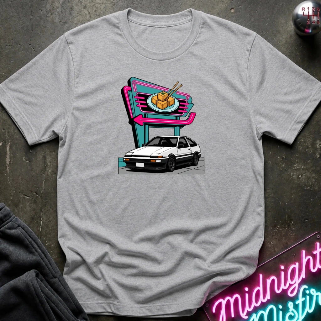 A light gray short-sleeve t-shirt laid flat on a dark textured surface, featuring a colorful graphic print of a white hatchback parked in front of a neon-style food sign. 
