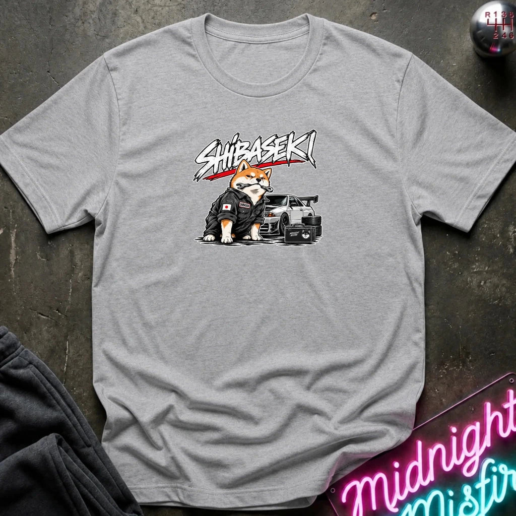 A light gray crewneck t-shirt laid flat on a dark textured surface. The shirt features a graphic print of a Shiba Inu dog wearing a black racing suit next to a black sports car, with the word 'SHIBASEKI' written above in a stylized white and red font.