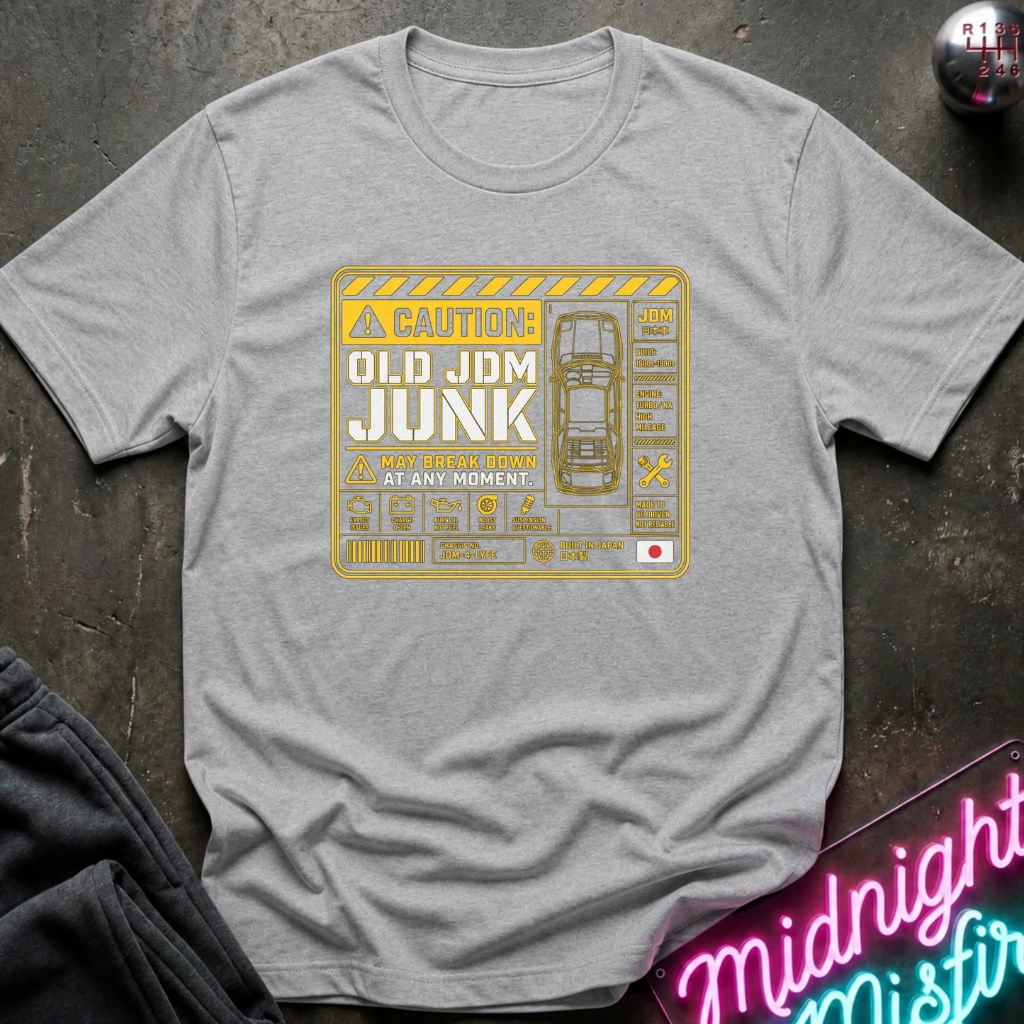 A light gray heather crewneck t-shirt with a yellow and white caution-style graphic reading 'OLD JDM JUNK' and 'A WAY BROKE DOWN AT ANY MOMENT', laid flat on a dark textured surface with partial jeans and a neon sign visible at the edges.