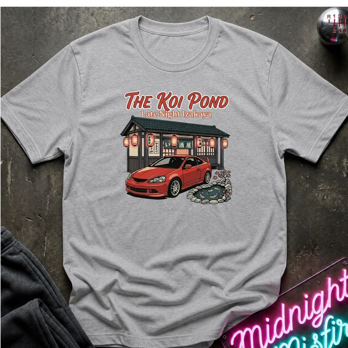 A light gray short-sleeve graphic t-shirt laid flat on a dark textured surface. The graphic features the text 'THE KOI POND Late Night Hamburger' above an illustration of a red sports car parked in front of a restaurant with red lanterns and a koi pond.lettering.