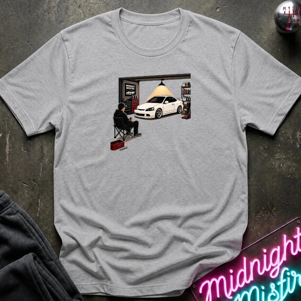 A light heather gray crewneck t-shirt laid flat on a dark textured surface. The shirt features a central graphic print of a person sitting in a folding chair in front of an open garage door, looking at a white sports car inside a garage.