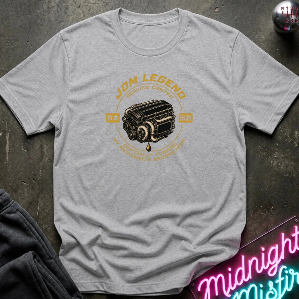 gray graphic t-shirt laid flat on a dark textured surface. The shirt features a vintage-style yellow and black engine graphic with the text 'JDM LEGEND' and 'MY SPECIALTY FUNDS MECHANIC'S SECOND HAND'.