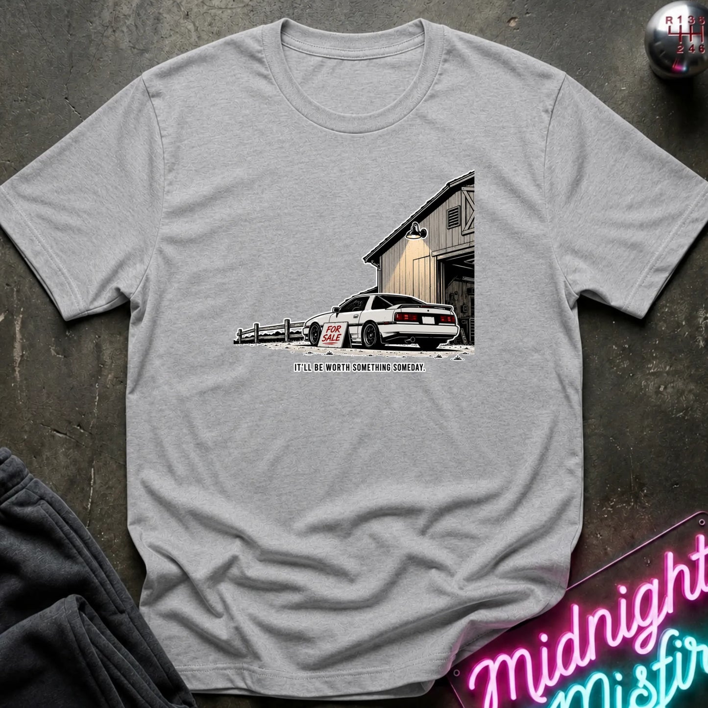 A light gray crewneck t-shirt laid flat on a dark textured surface, featuring a graphic print of a white sports car parked in front of a barn with the text.