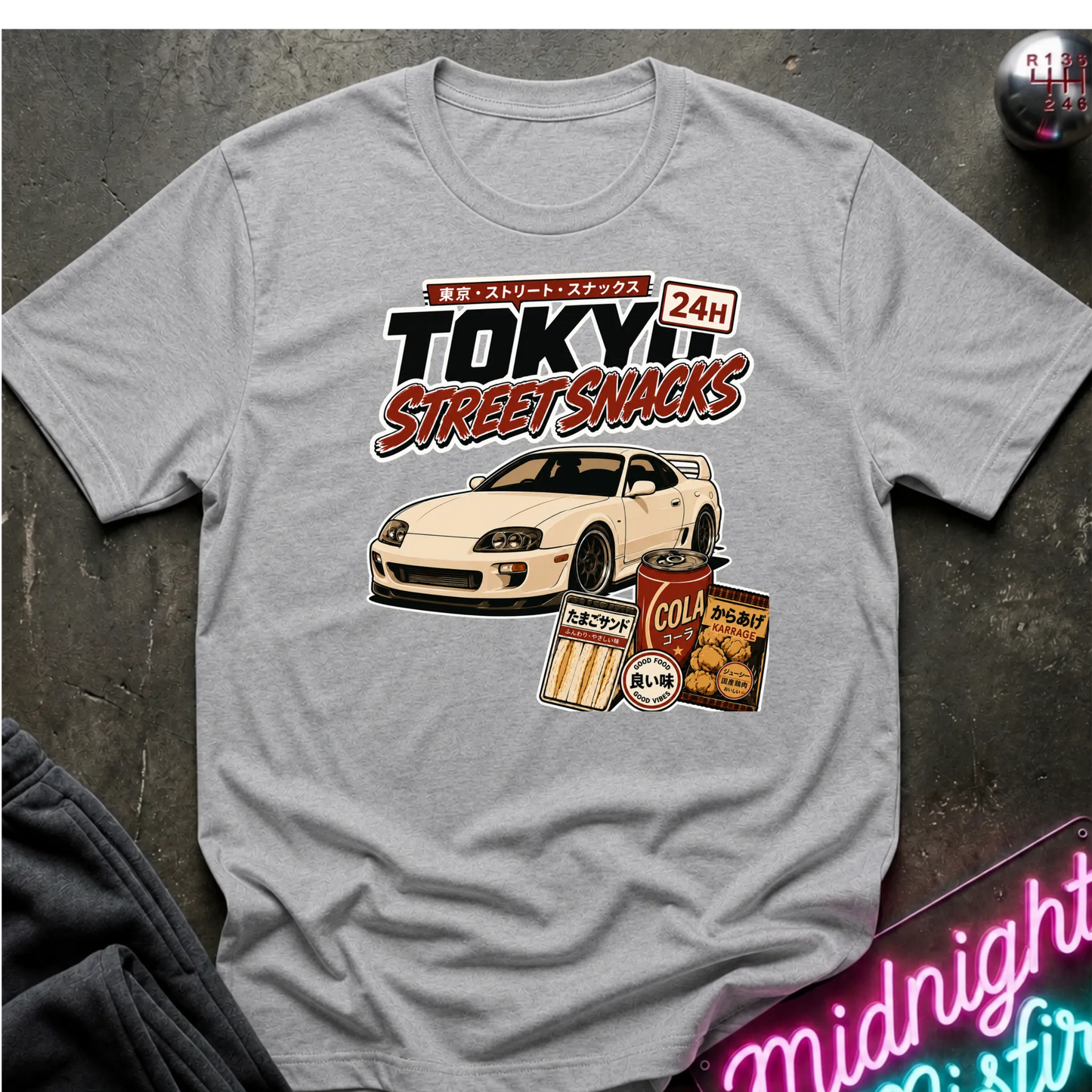 A light gray graphic t-shirt laid flat on a dark textured surface. The graphic features a white sports car and retro Japanese-style snack cans with the text 'TOKYO STREET SNACKS 24H'.