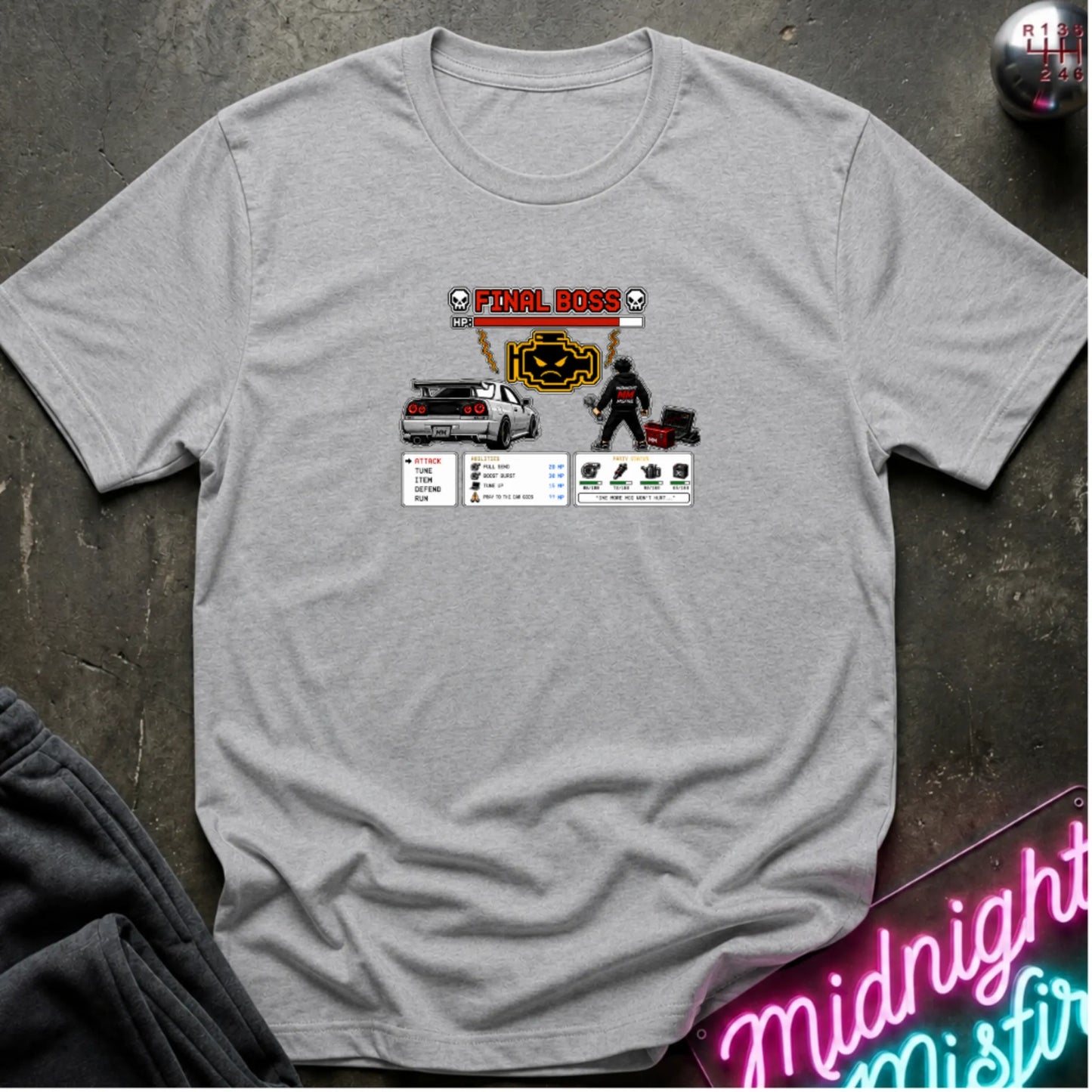 A heather gray short-sleeve crewneck t-shirt laid flat on a dark textured surface, featuring a central graphic print of a rpg character and a car. 