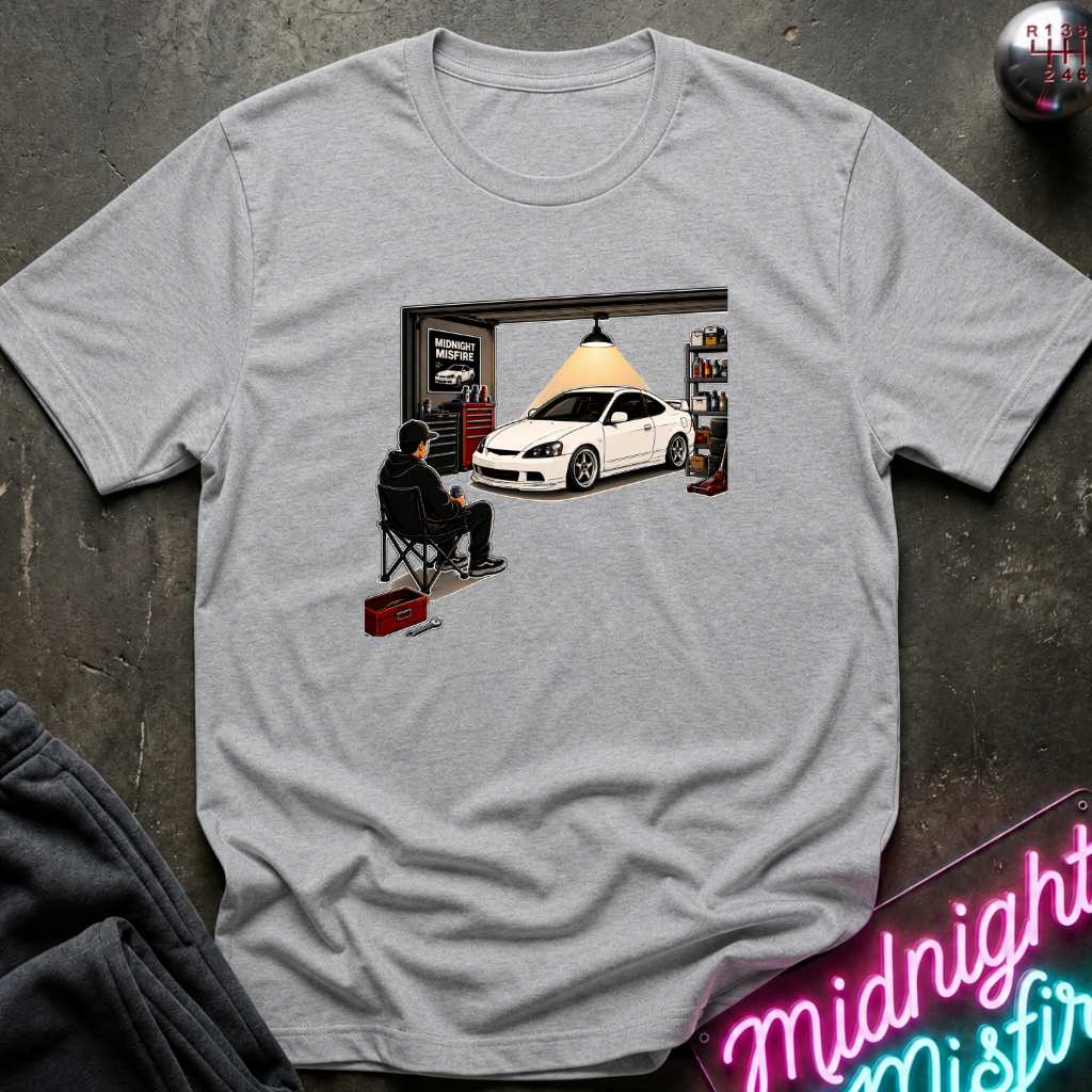 A light gray crewneck t-shirt featuring a graphic print of a man sitting in a folding chair in front of an open garage door with a white sports car inside. The shirt is displayed flat on a dark textured surface with a neon sign and dark sweatpants partially visible.