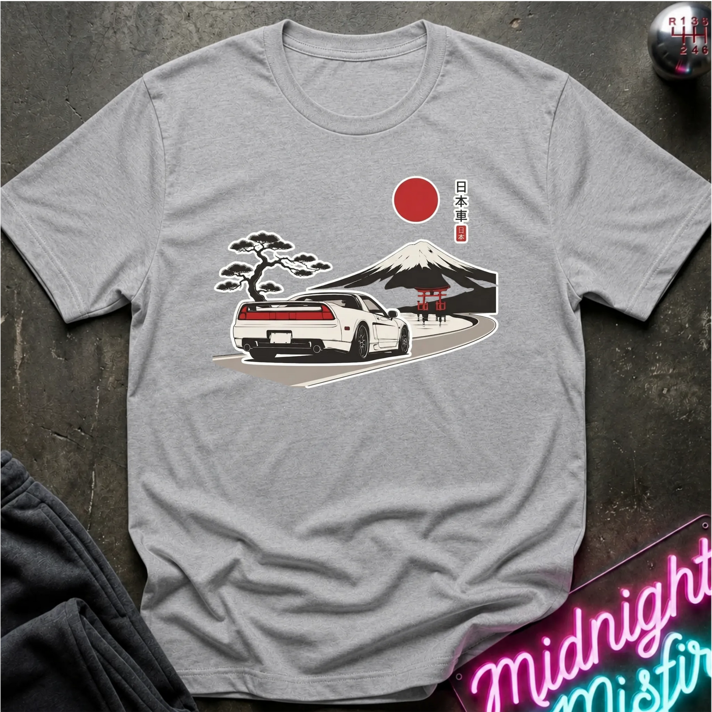 A light gray crewneck t-shirt featuring a graphic print of a white sports car on a road with Mount Fuji, a cherry blossom tree, a red sun, and Japanese text. The shirt is laid flat on a dark textured surface with a neon sign and dark pants partially visible.