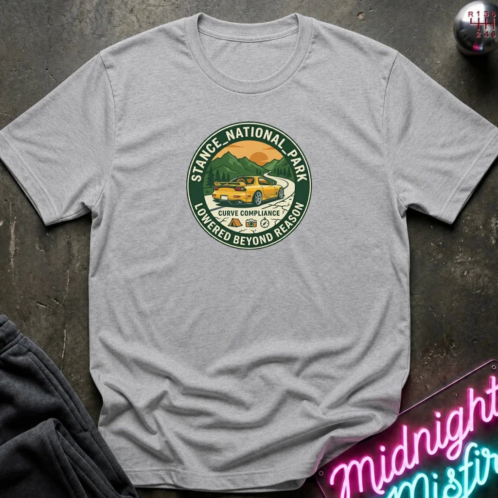 A light heather gray short-sleeve t-shirt laid flat on a dark textured surface, featuring a circular graphic print of a yellow car in a scenic mountain landscape with the text 'STACE NATIONAL PARK' and 'LOWEDGED BEYOND REASON'.