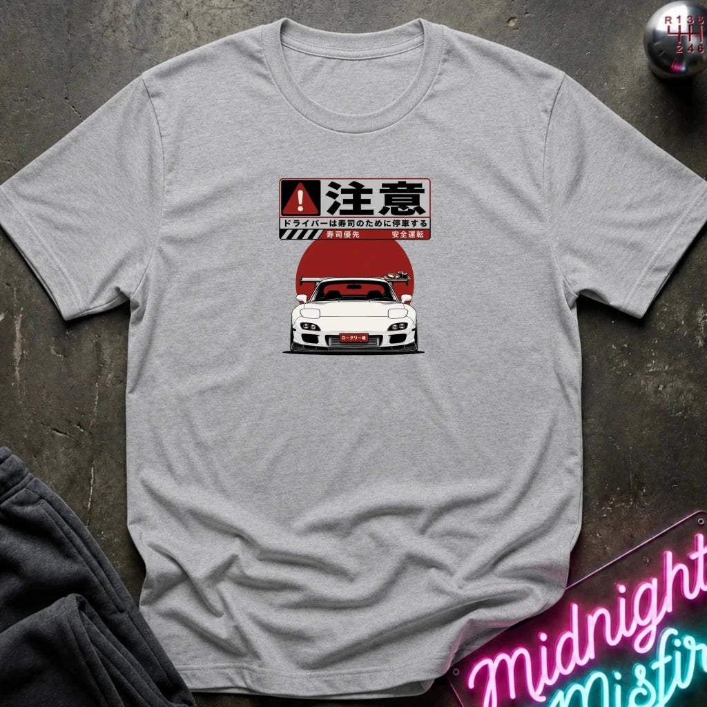 A light gray crewneck t-shirt laid flat on a dark textured surface, featuring a graphic print of a red and white sports car with Japanese text and a warning sign motif. 