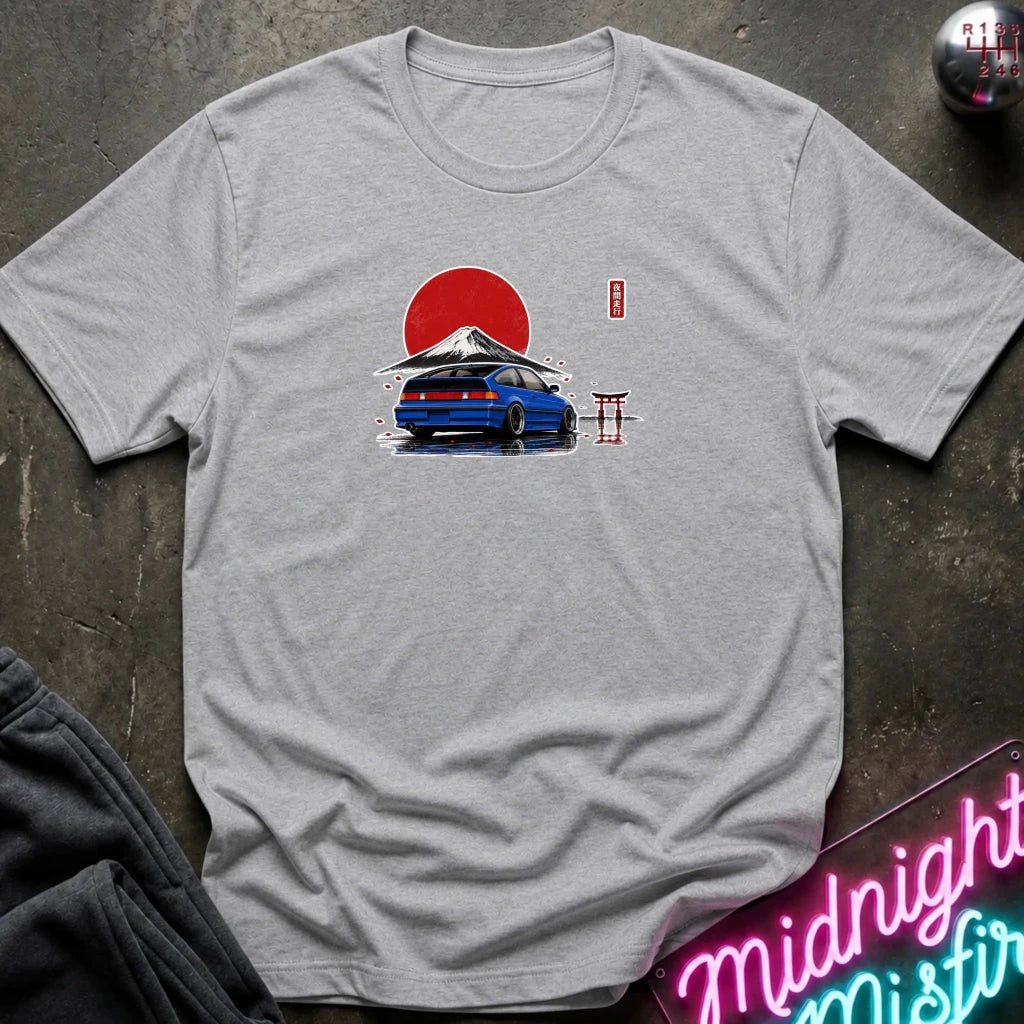 A heather gray crewneck t-shirt featuring a graphic print of a blue station wagon parked in front of a red Japanese sun with Mount Fuji and a torii gate. The shirt is displayed flat on a dark textured surface with a neon sign and sweatpants partially visible.