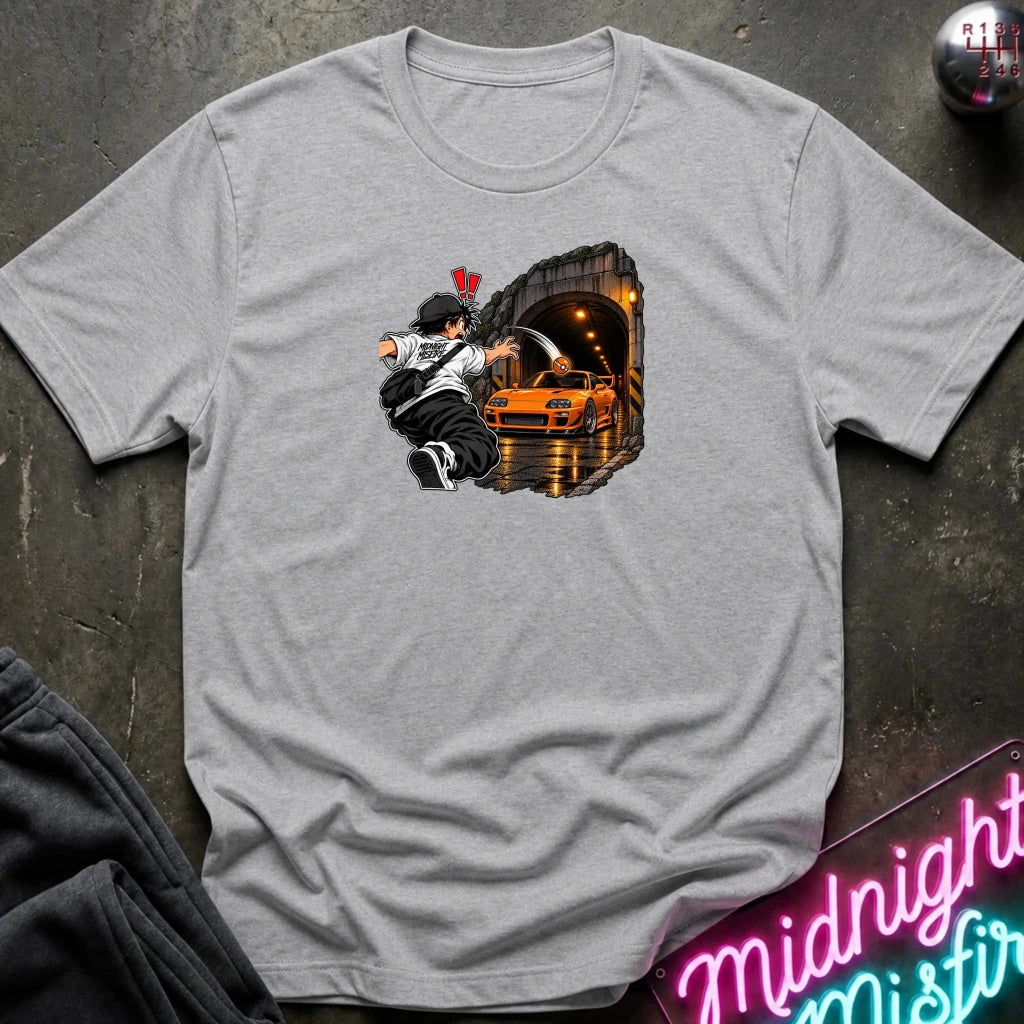 A light gray short-sleeve crewneck t-shirt laid flat on a dark textured surface. The center of the shirt features a graphic print of an anime character in a black and white outfit with a hat, next to a sports car 