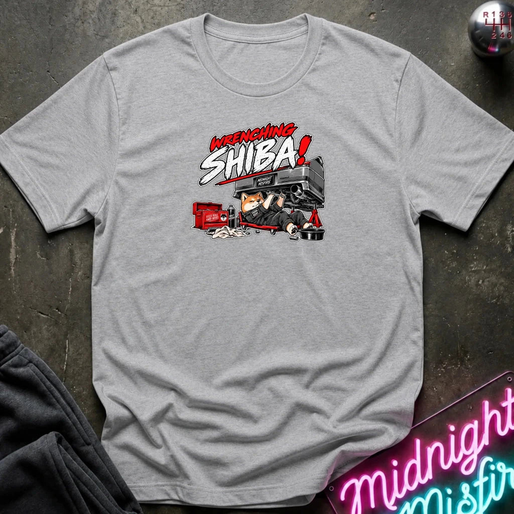 A light gray heather short-sleeve graphic t-shirt laid flat on a dark textured surface. The graphic features the text 'WRENCHING SHIBA!' in red and white comic-style lettering above an illustration of a character in a black and red vehicle.