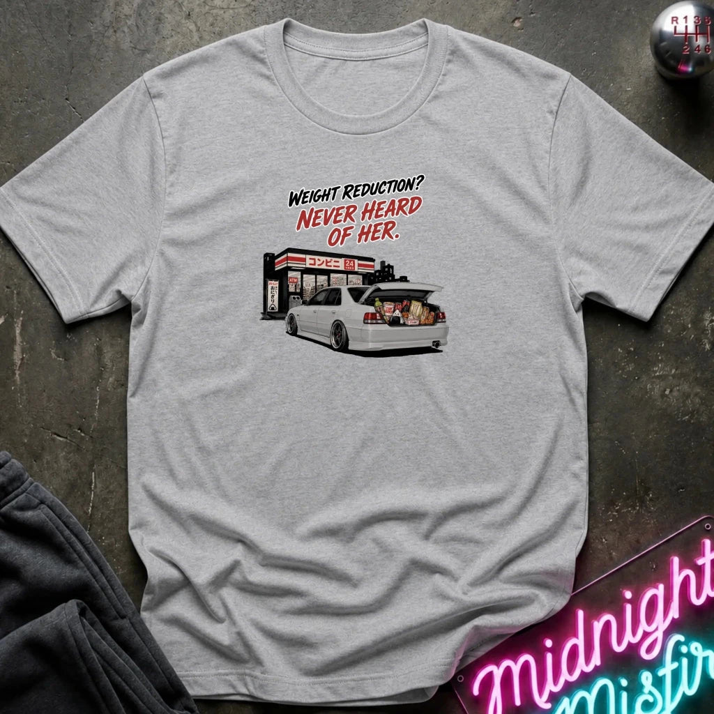 A light gray heather crewneck t-shirt laid flat on a dark textured surface. The shirt features a graphic print of a white station wagon with its trunk open, parked in front of a convenience store, with the text 'WEIGHT REDUCTION? NEVER HEARD OF HER.