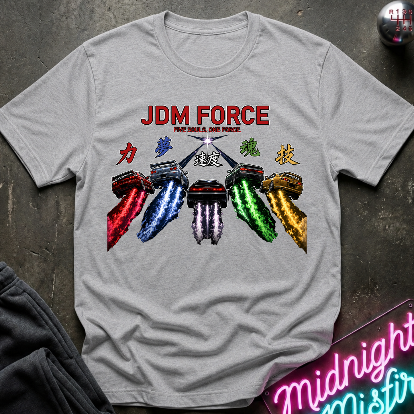 A light gray crewneck t-shirt featuring a colorful graphic print of Japanese motorcycle racing cars with the text 'JDM FORCE'. The shirt is displayed flat on a dark textured surface with lifestyle props like a neon sign and a pair of dark sweatpants.