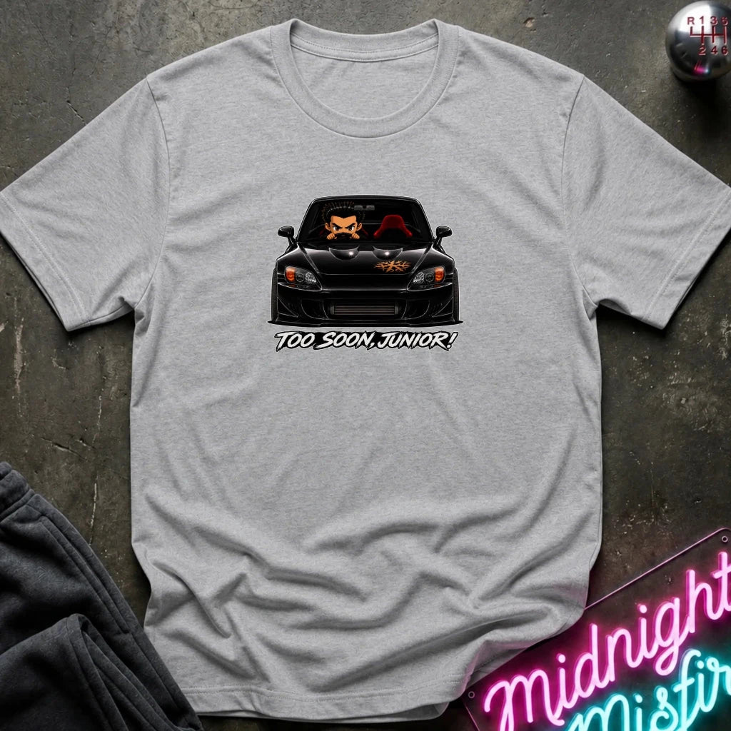 A light heather gray t-shirt laid flat on a dark textured surface, featuring a graphic of a black sports car with orange cat faces in the windshield and the text 'TOO SOON, JUNIOR!' printed below. A neon sign and dark clothing are partially visible in the background as props.