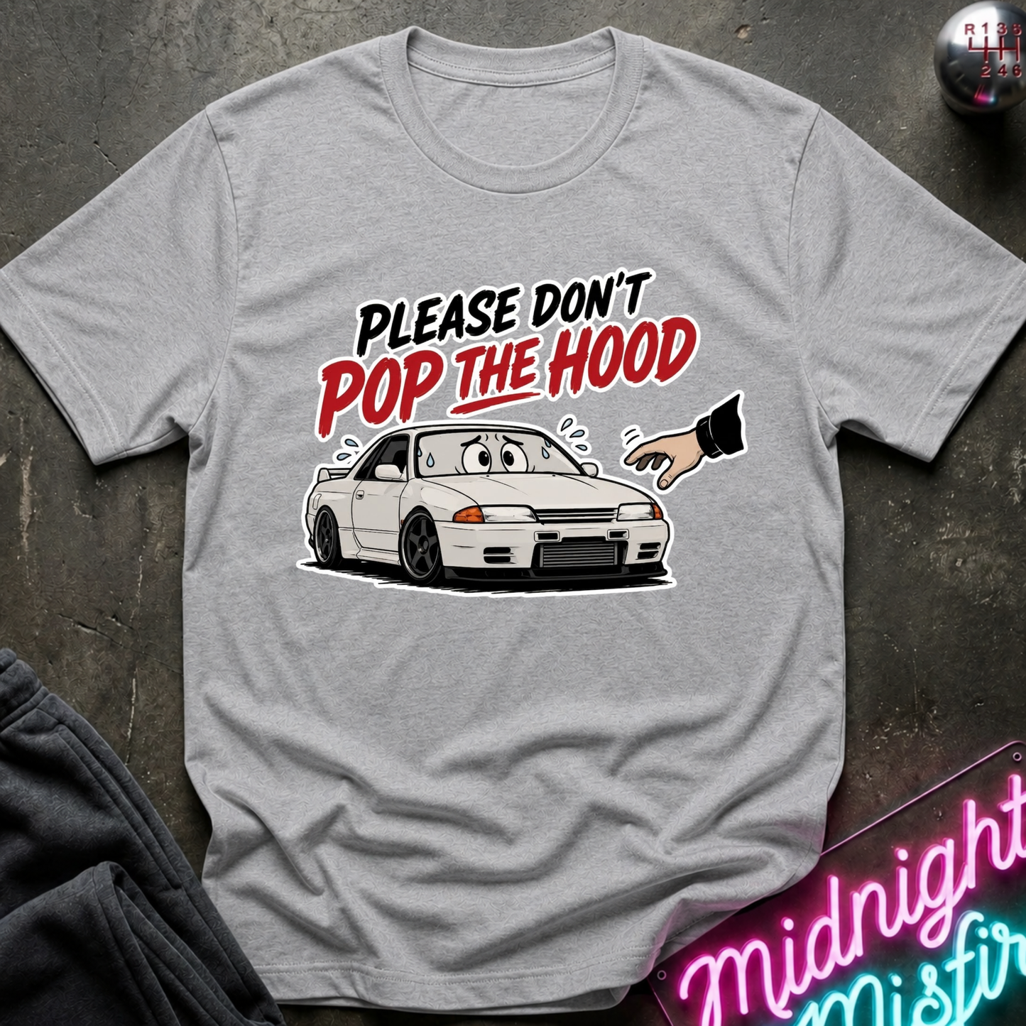 A light gray heather crewneck t-shirt featuring a graphic of a white Nissan R32 GT-R with the text 'PLEASE DON'T POP THE HOOD' in black and red. The shirt is displayed flat on a dark textured surface with a neon sign and dark pants partially visible.