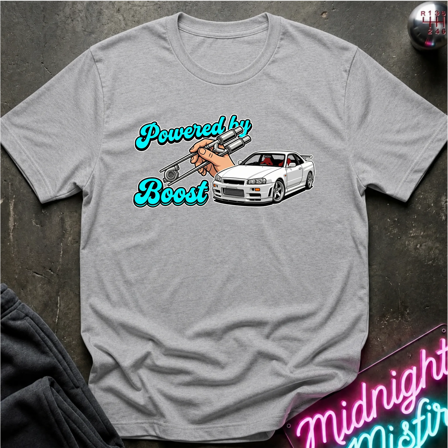 A light gray heather t-shirt laid flat on a dark textured surface, featuring a graphic print of a white sports car, a hand holding a silver boost gauge, and the text 'Powered by Boost' in a bright teal script.