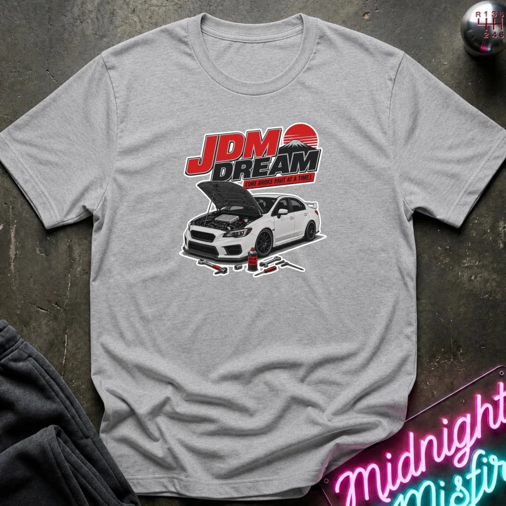 A heather gray short-sleeve t-shirt laid flat on a dark textured surface, featuring a graphic print of a white sports car with its hood open beneath the text 'JDM DREAM' in red and black. 
