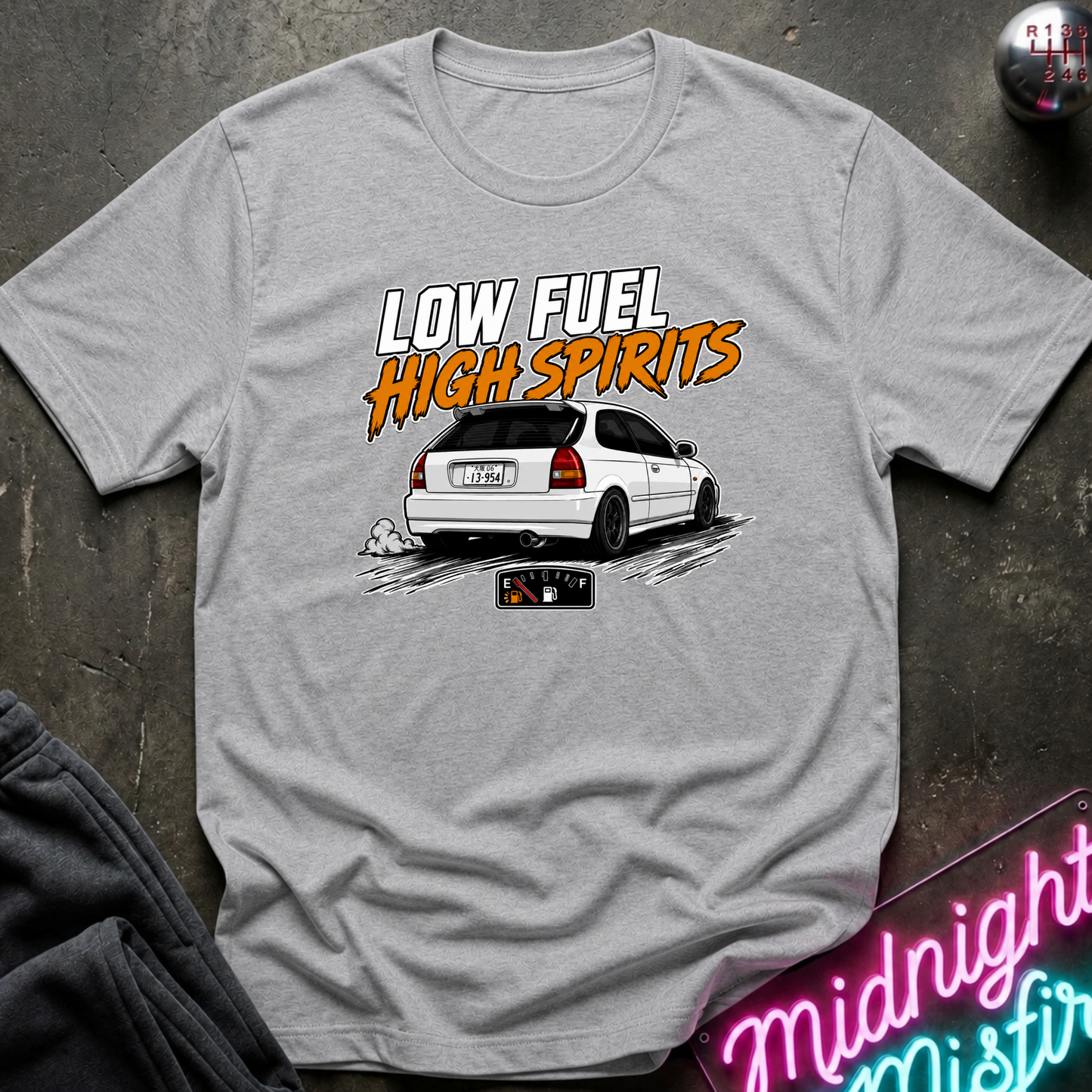 A light gray heather t-shirt laid flat on a dark textured surface, featuring a graphic print of a white hatchback car with the text 'LOW FUEL HIGH SPIRITS' in white and orange. A neon sign and dark clothing are partially visible in the styled flat-lay composition.