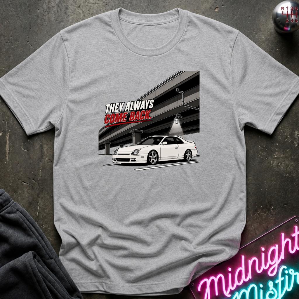 A light gray heather crewneck t-shirt laid flat on a dark textured surface. The shirt features a graphic print of a white sports car parked under a highway structure, with the text 'THEY ALWAYS COME BACK' in white and red.