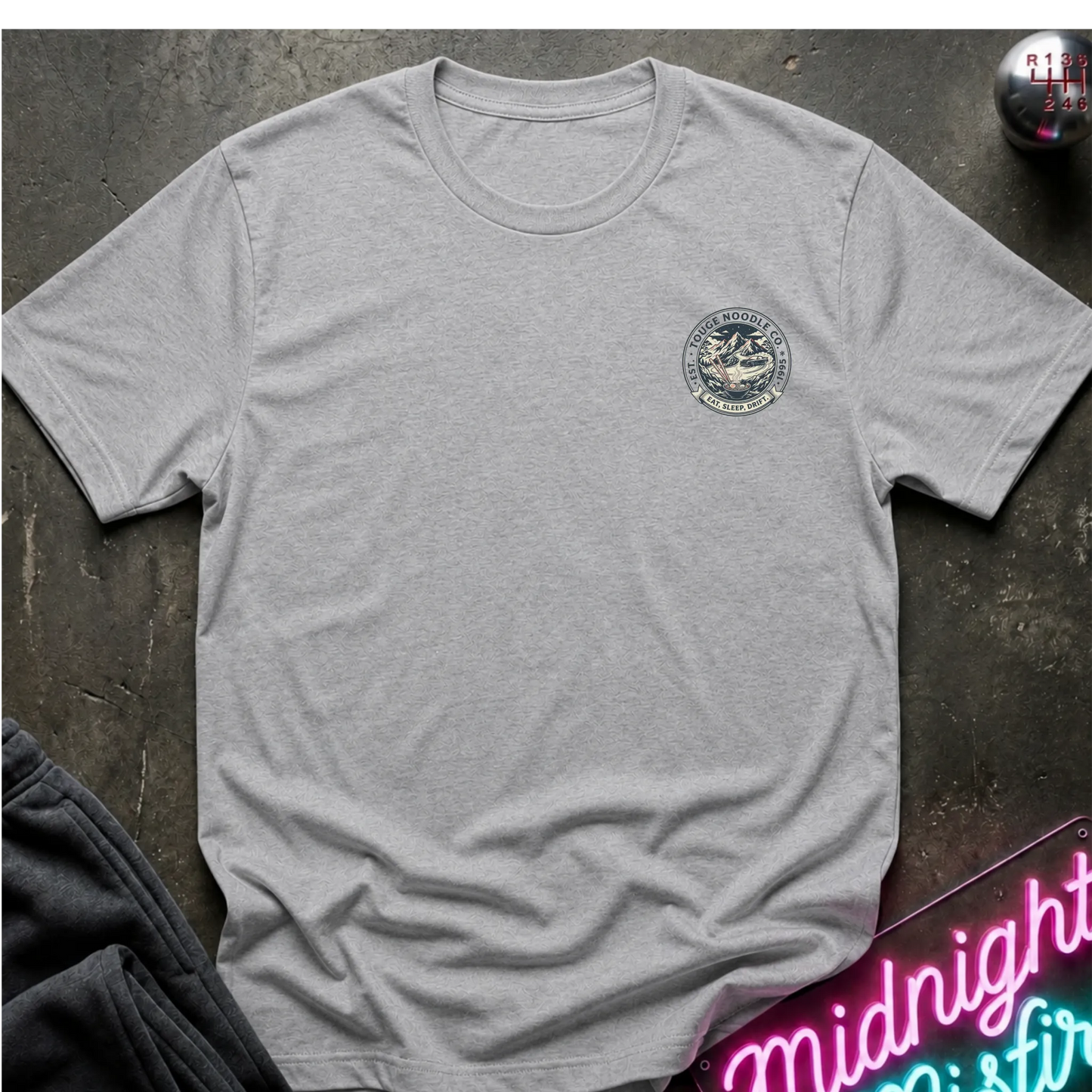 Gray crewneck t-shirt with a circular crest printed on the left chest, styled flat on a dark textured surface with a neon sign and dark pants in the background.