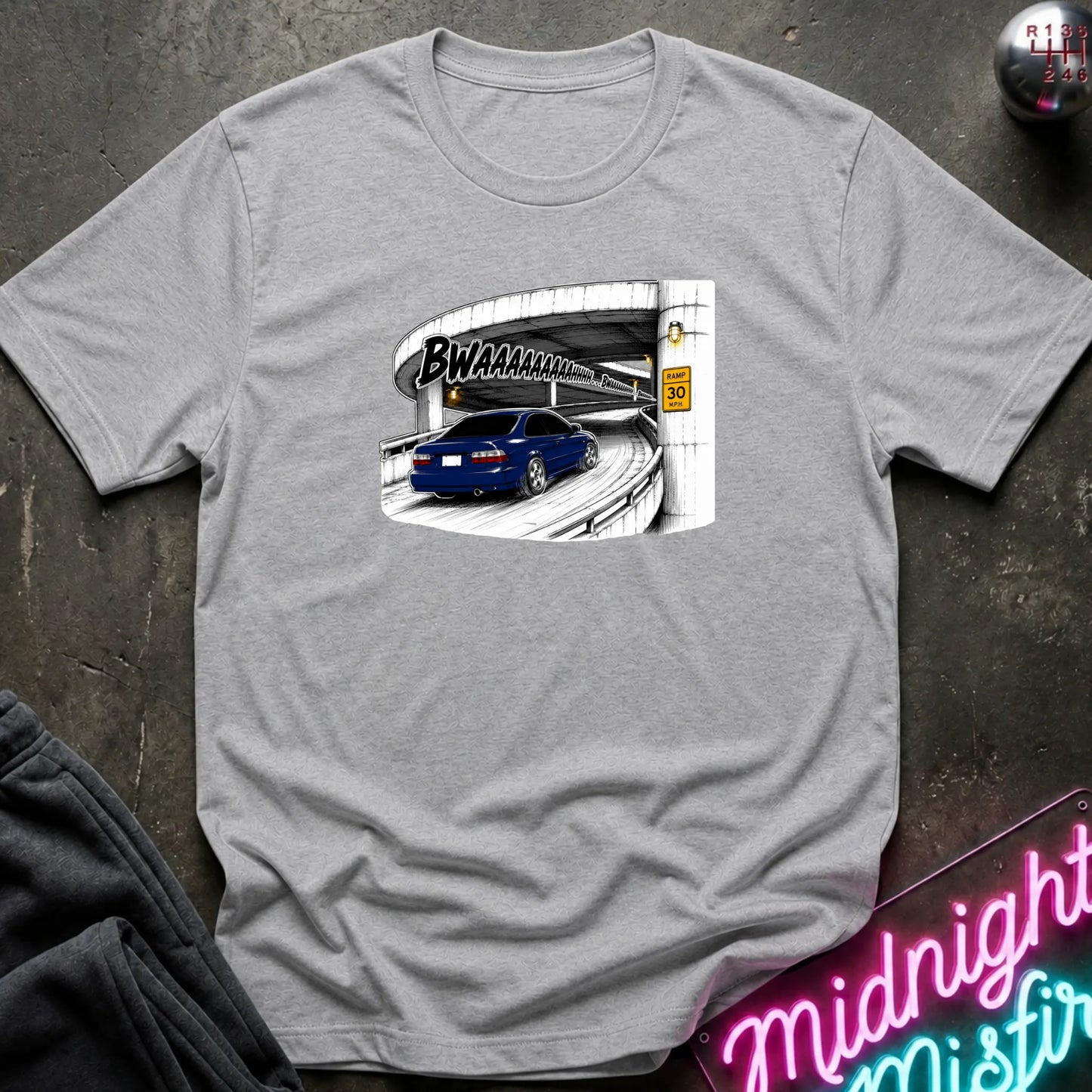 A light gray short-sleeve crewneck t-shirt laid flat on a dark textured surface. The shirt features a graphic print of a blue car driving under a highway overpass with the text 'BWAHAHA'.