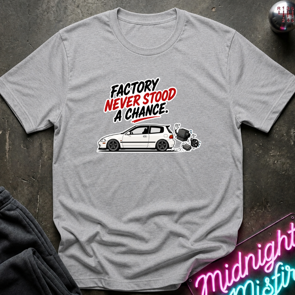 A heather gray short-sleeve t-shirt laid flat on a dark textured surface. The shirt features a graphic print with the text 'FACTORY NEVER STOOD A CHANCE' in black and red, above a white car and a black-and-white dog.