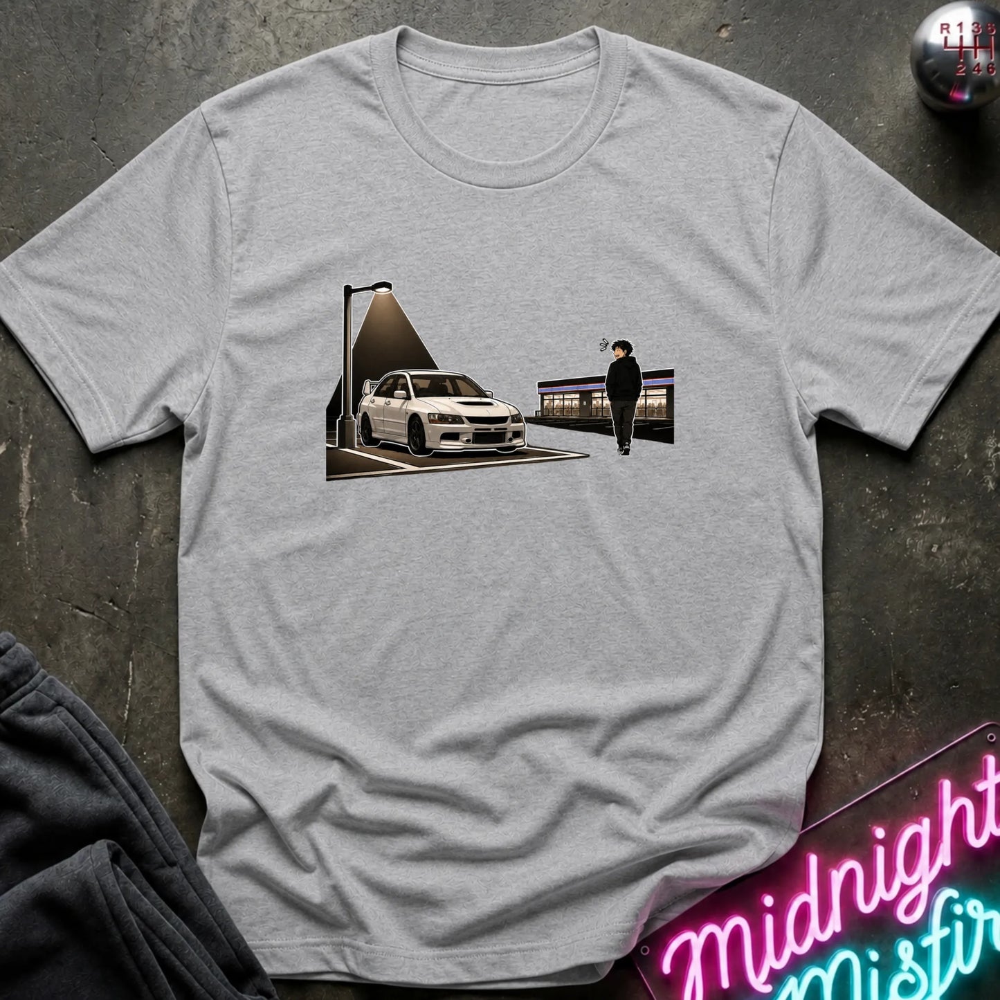 A light gray heather crewneck t-shirt featuring a graphic print of a white sports car and a person under a streetlamp, laid flat on a dark textured surface with a neon sign and dark pants partially visible.