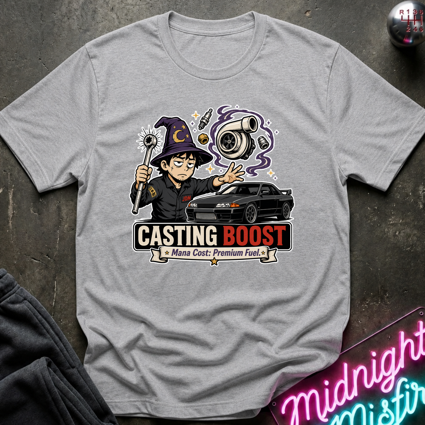 A light gray crewneck t-shirt featuring a graphic print of a cartoon wizard casting a spell next to a black sports car, with the text 'CASTING BOOST' and 'Mana Cost: Premium Fuel'. The shirt is displayed flat on a dark textured surface with a neon sign and dark fabric in the corners.