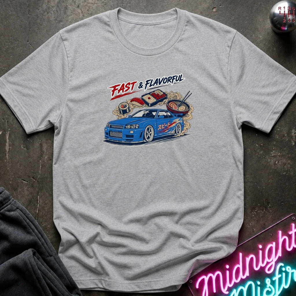 A light gray heather short-sleeve graphic T-shirt laid flat on a dark textured surface. The shirt features a colorful print of a blue sports car with ramen noodles and chopsticks, plus the text 'FAST & FLAVORFUL' in red and black.