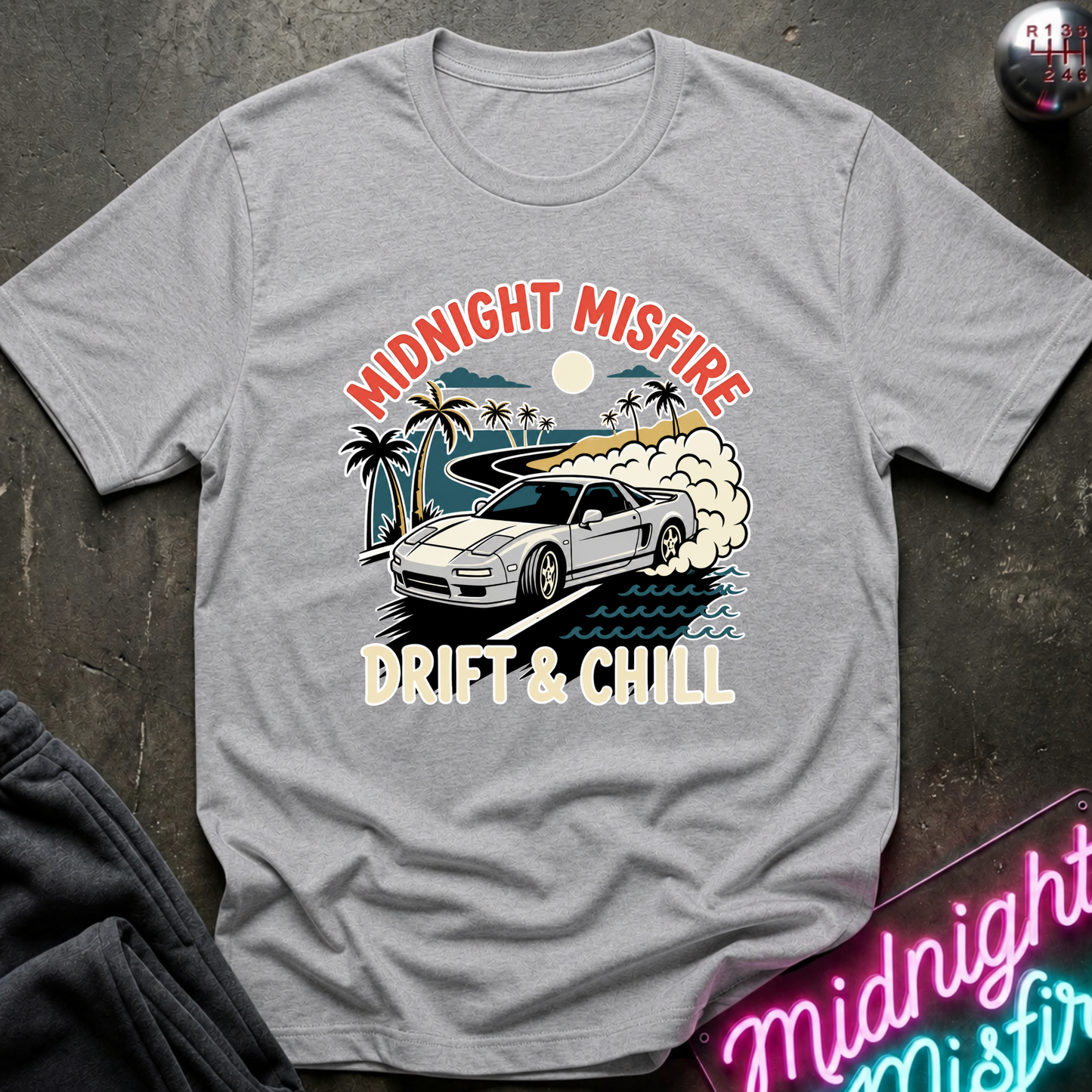 A light gray heather t-shirt with a graphic print of a white sports car drifting on a beach with palm trees and the text 'MIDNIGHT MISFIRE DRIFT & CHILL'. The t-shirt is laid flat on a dark textured surface with a neon sign and dark pants partially visible.