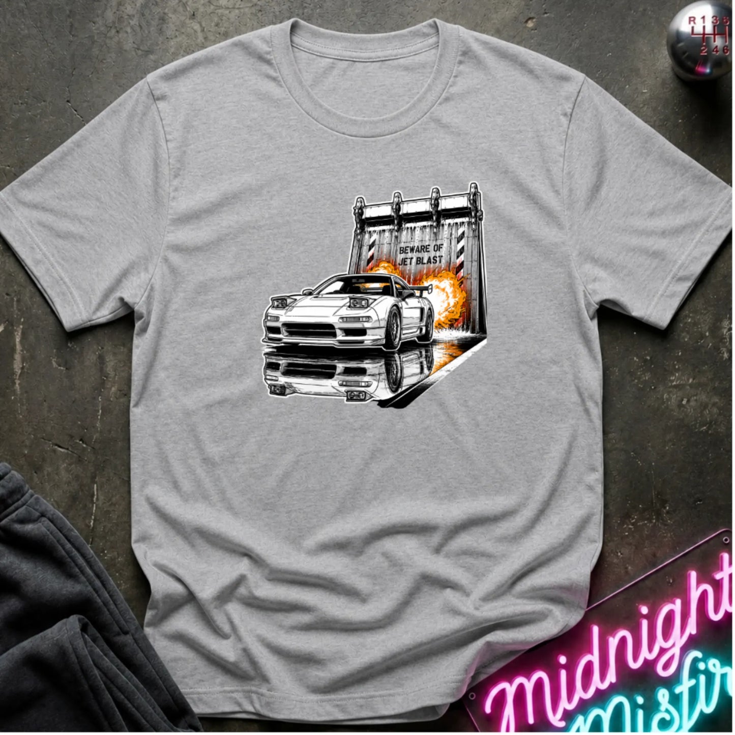 A heather gray short-sleeve t-shirt laid flat on a dark textured surface, featuring a graphic print of a white sports car in front of a blast off area.