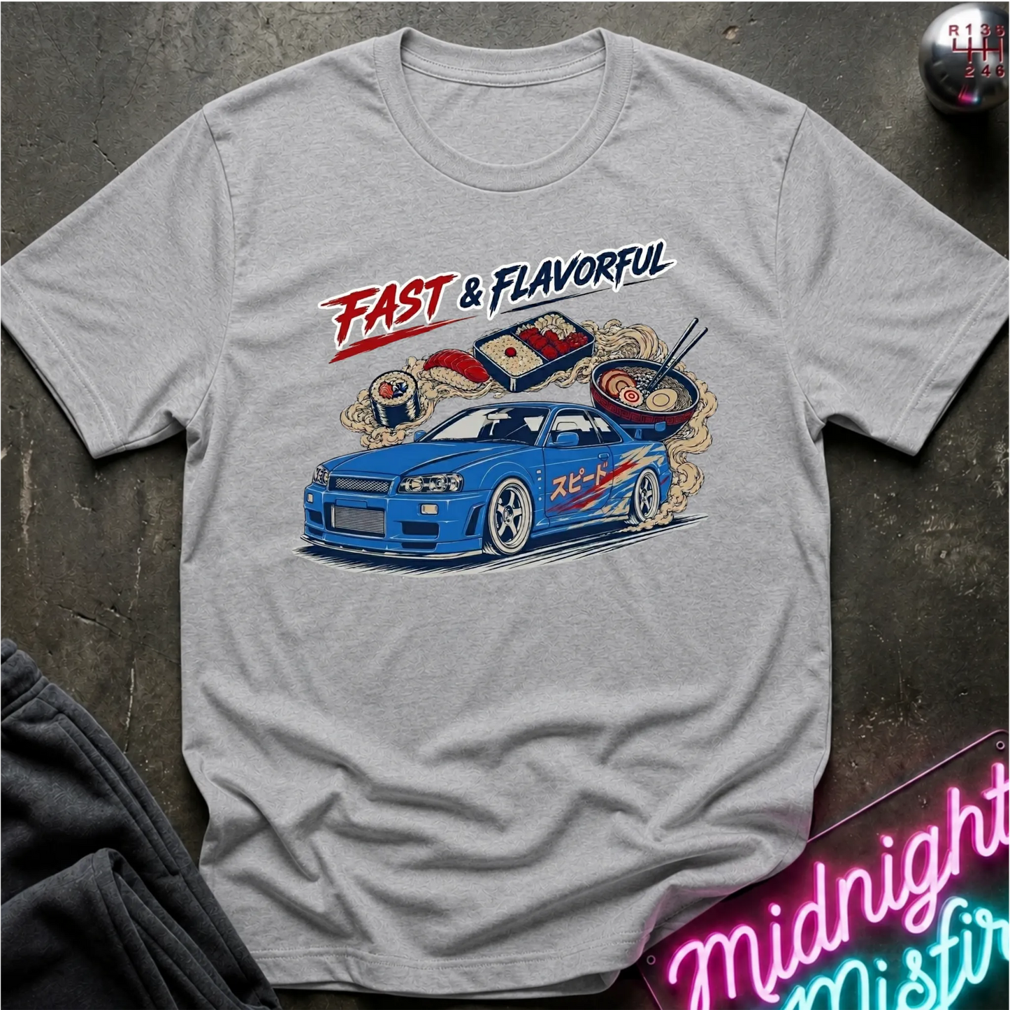 A light gray crewneck t-shirt featuring a graphic print of a blue sports car with sushi-themed details and the text 'FAST & FLAVORFUL'. The shirt is laid flat on a dark textured surface with a neon sign and dark fabric partially visible in the corners.