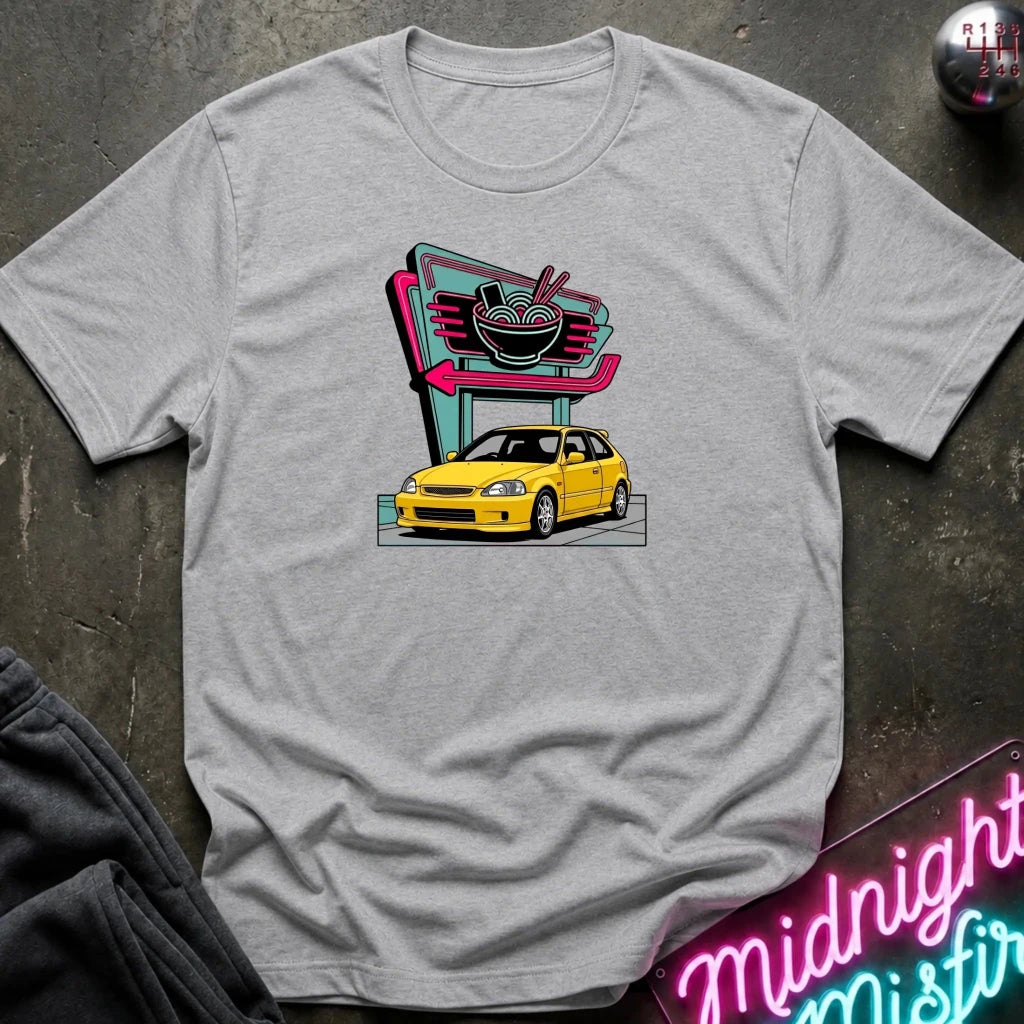 A light heather gray short-sleeve t-shirt laid flat on a dark textured surface, featuring a retro-style graphic of a yellow hatchback car parked in front of a neon ramen shop sign. 