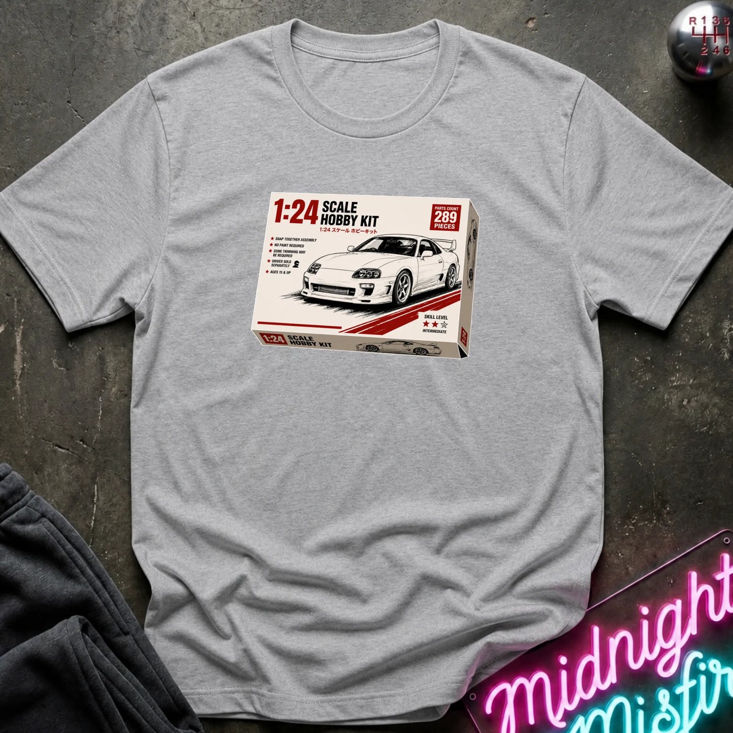 A light gray heather crewneck t-shirt laid flat on a dark textured surface, featuring a graphic print of a 1:24 scale model kit box for a white sports car.