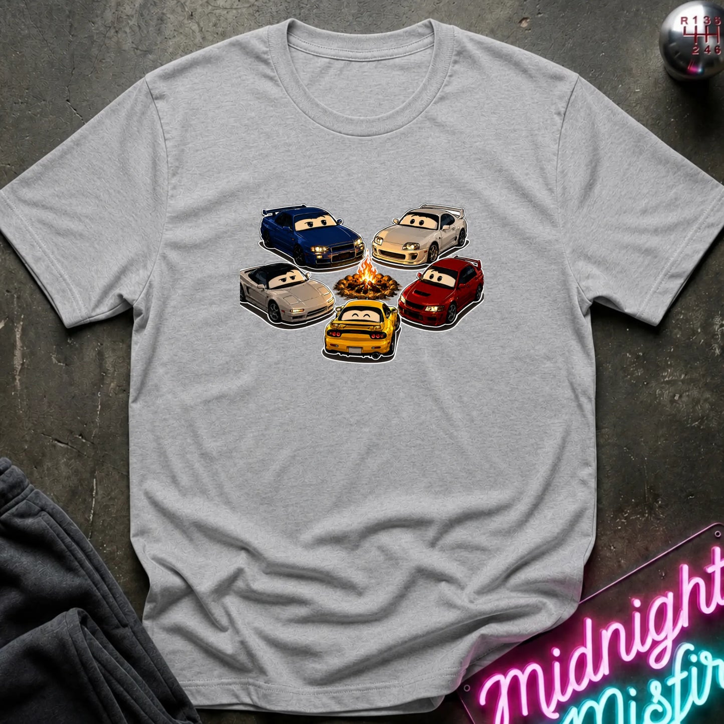 A light gray heather short-sleeve t-shirt laid flat on a dark textured surface, featuring a graphic of cartoon race cars arranged around a campfire. The shirt is styled with a gray sweatshirt in the corner and a pink neon sign in the bottom right.