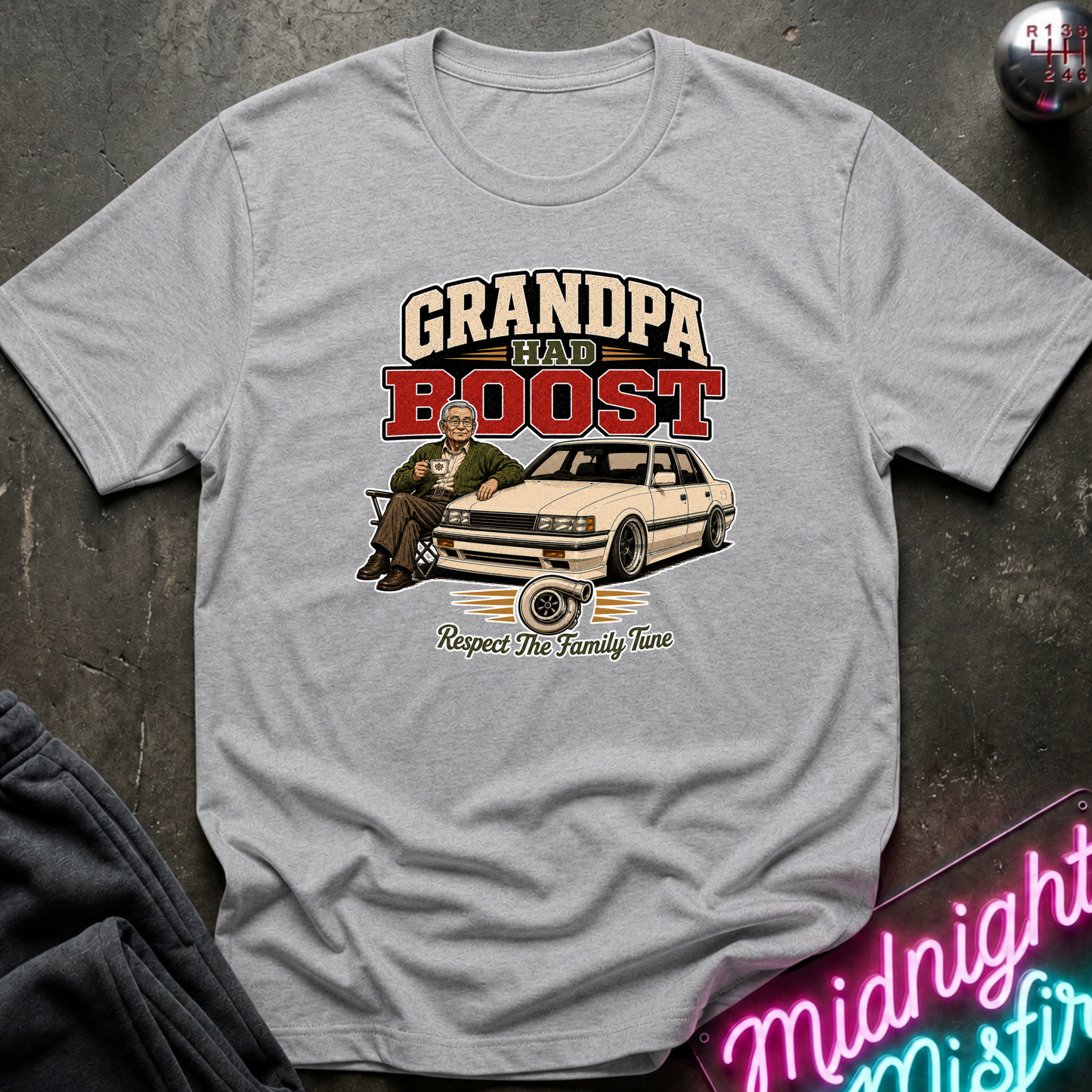A light gray heather crewneck t-shirt featuring a vintage-style graphic of a man sitting on a car with the text 'GRANDPA HAD A BOOST' and 'Respect The Family Tune'. The shirt is displayed flat on a dark textured surface with a neon sign and dark pants partially visible.