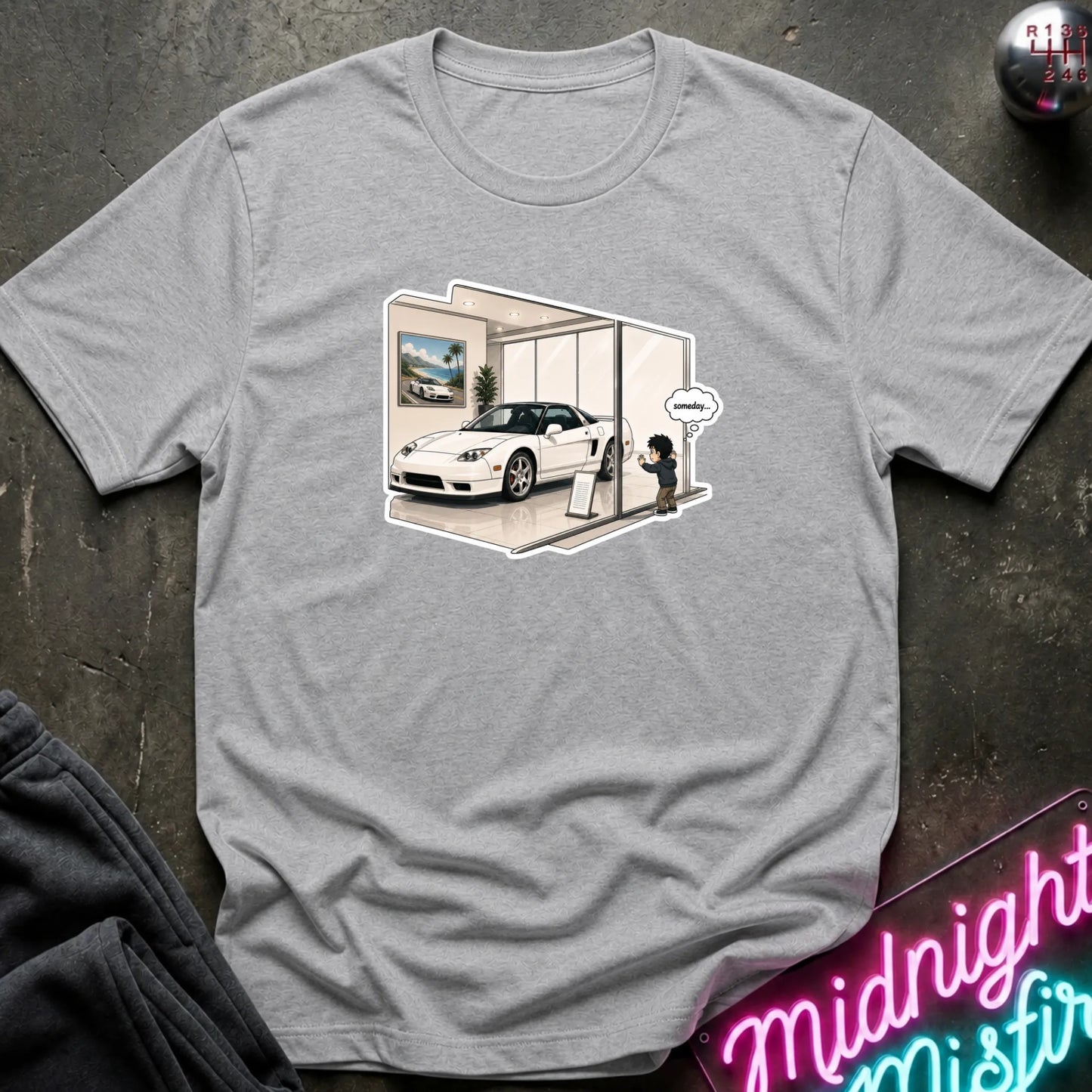 A heather gray short-sleeve t-shirt laid flat on a dark textured surface, featuring a cartoon graphic of a white sports car inside a glass showroom with a small character standing outside it. A neon sign is partially visible in the bottom right corner.