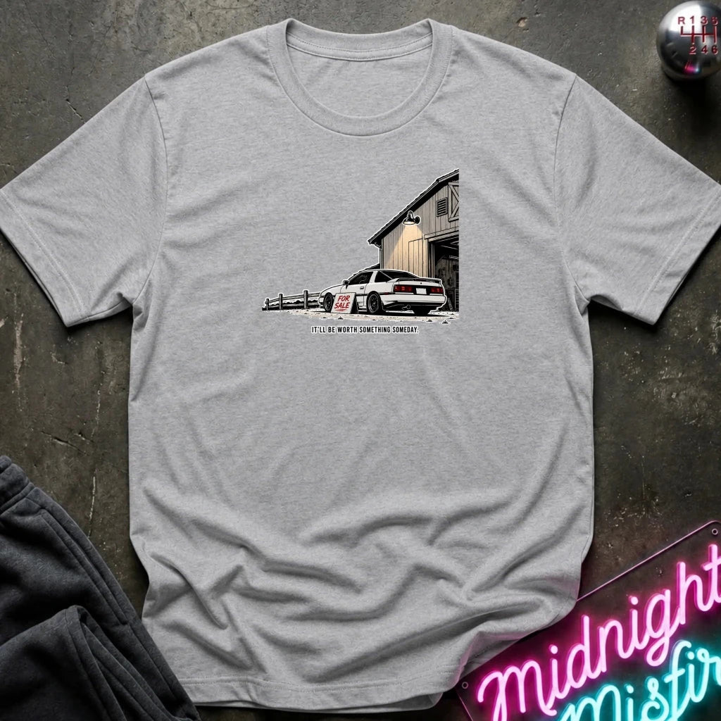 A light gray heather crewneck t-shirt laid flat on a dark textured surface. The shirt features a graphic print of a white muscle car parked in front of a barn 