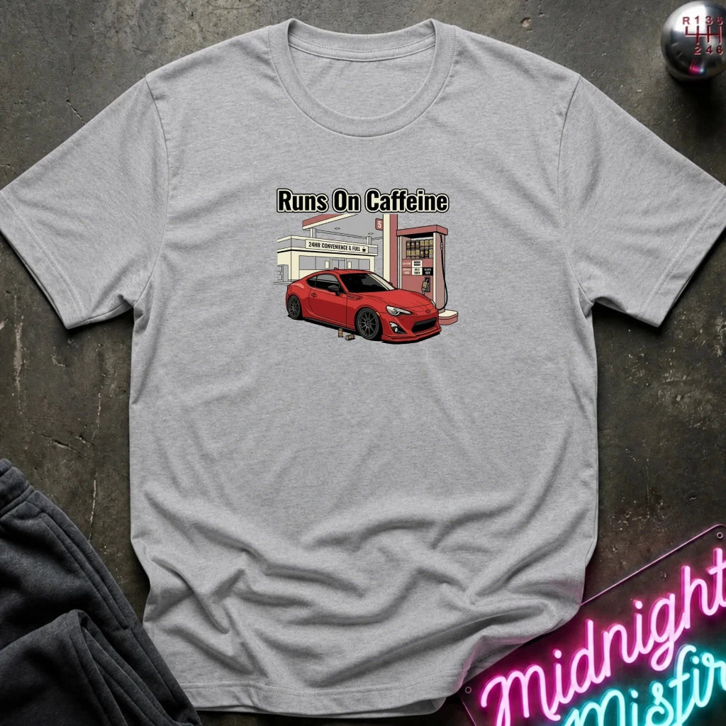 A heather gray crewneck t-shirt featuring a graphic of a red sports car at a vintage gas station with the text 'Runs On Caffeine'. The shirt is displayed flat on a dark textured surface with a neon sign and dark pants partially visible.